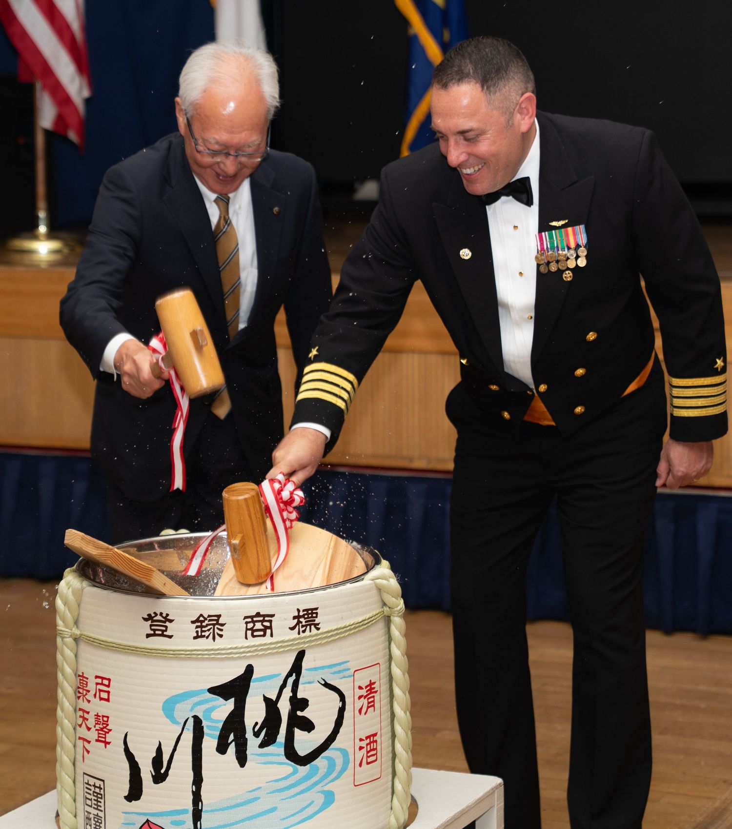 Two men in formal attire perform traditional Japanese sake barrel breaking ceremony at official military diplomatic event.