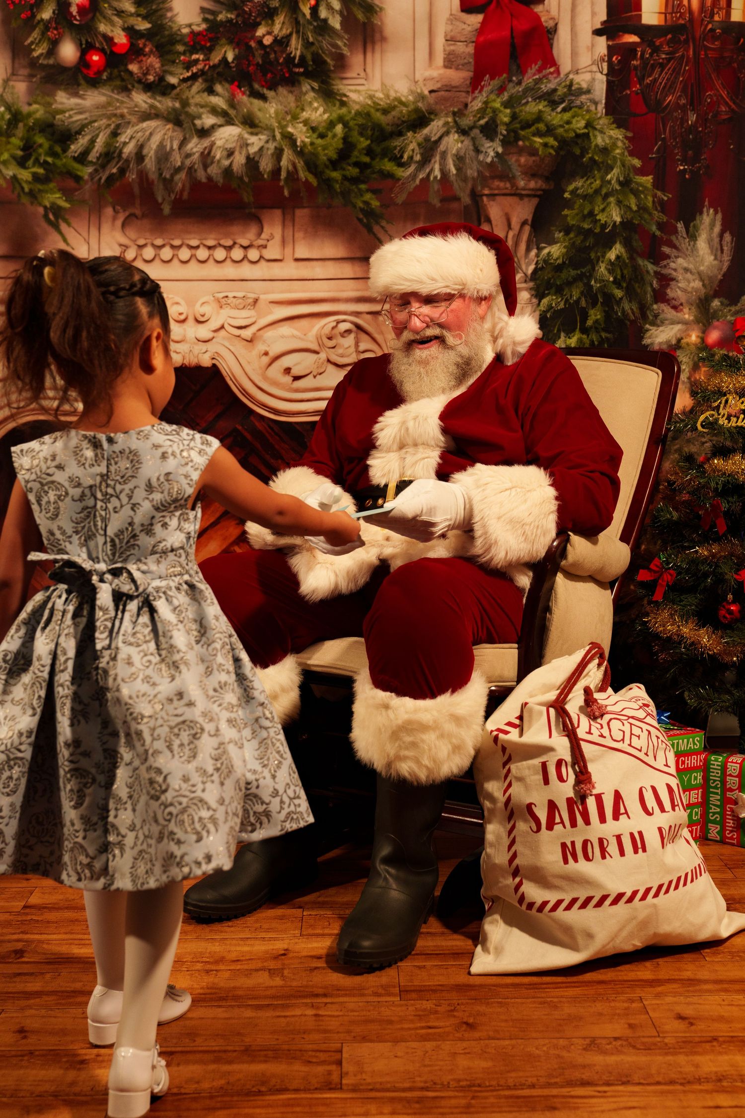 Young girl in silver dress visits Santa Claus seated in festive Christmas decorated setting with holiday gifts.