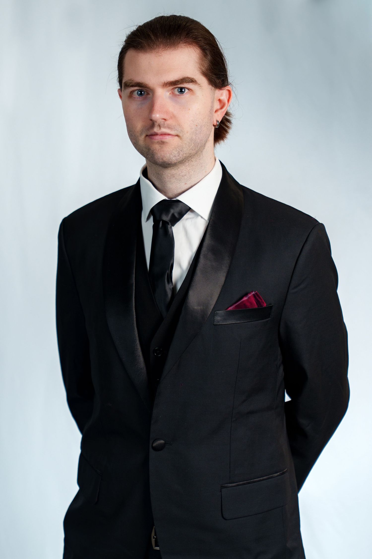 Professional man in black tuxedo with red pocket square poses against light gray background.