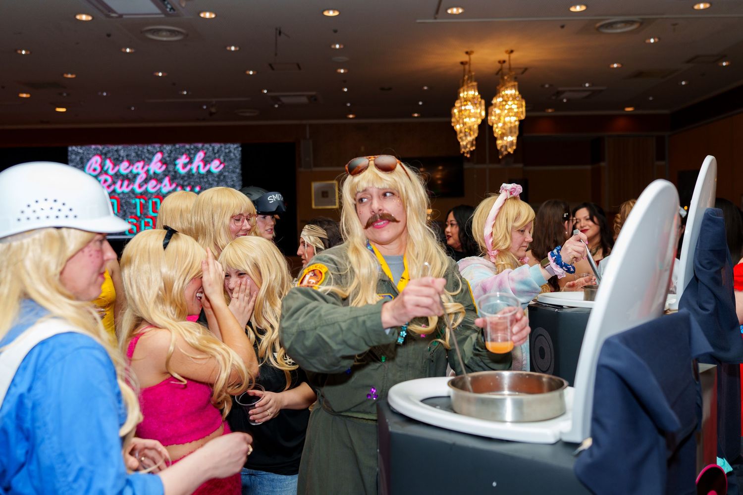Group of party guests wearing blonde wigs and costumes at a themed costume party in a hotel ballroom.