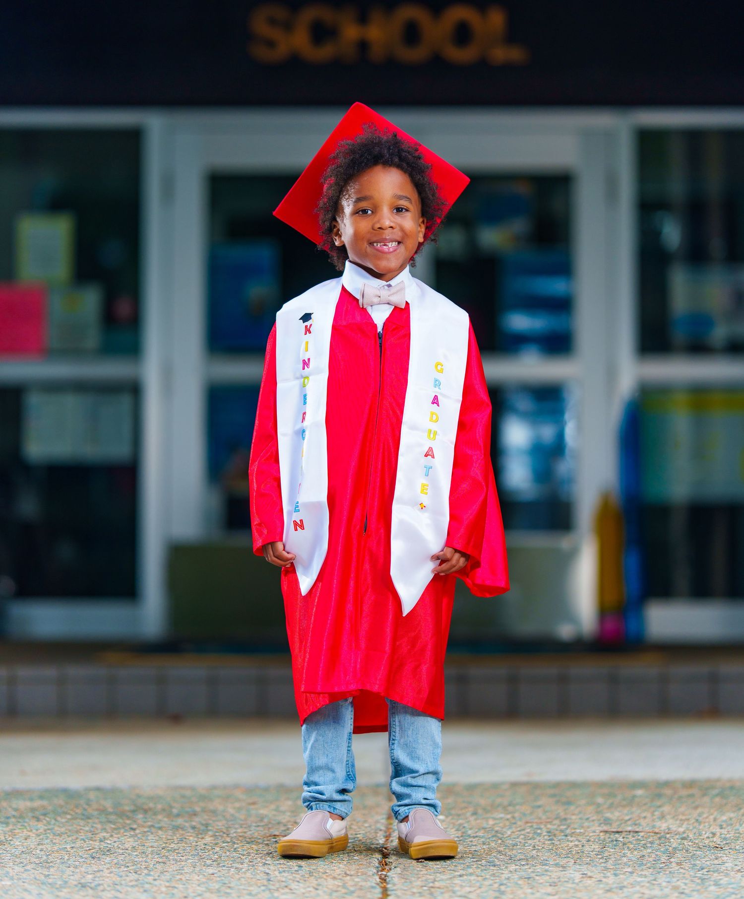 Young child in red graduation cap and gown smiling proudly in front of school entrance.