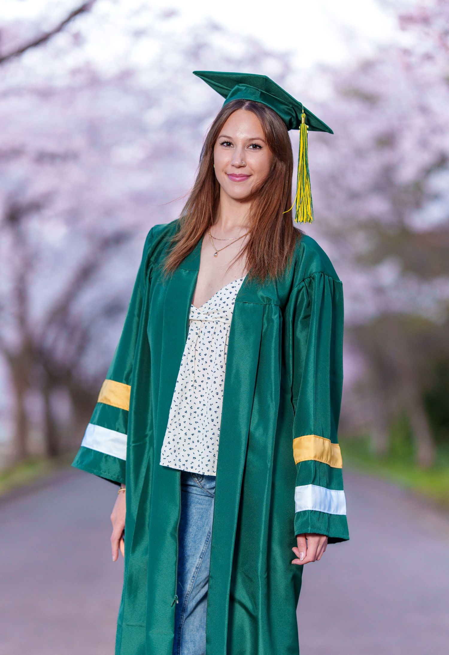 Smiling female graduate in green cap and gown stands on tree-lined path with cherry blossoms.