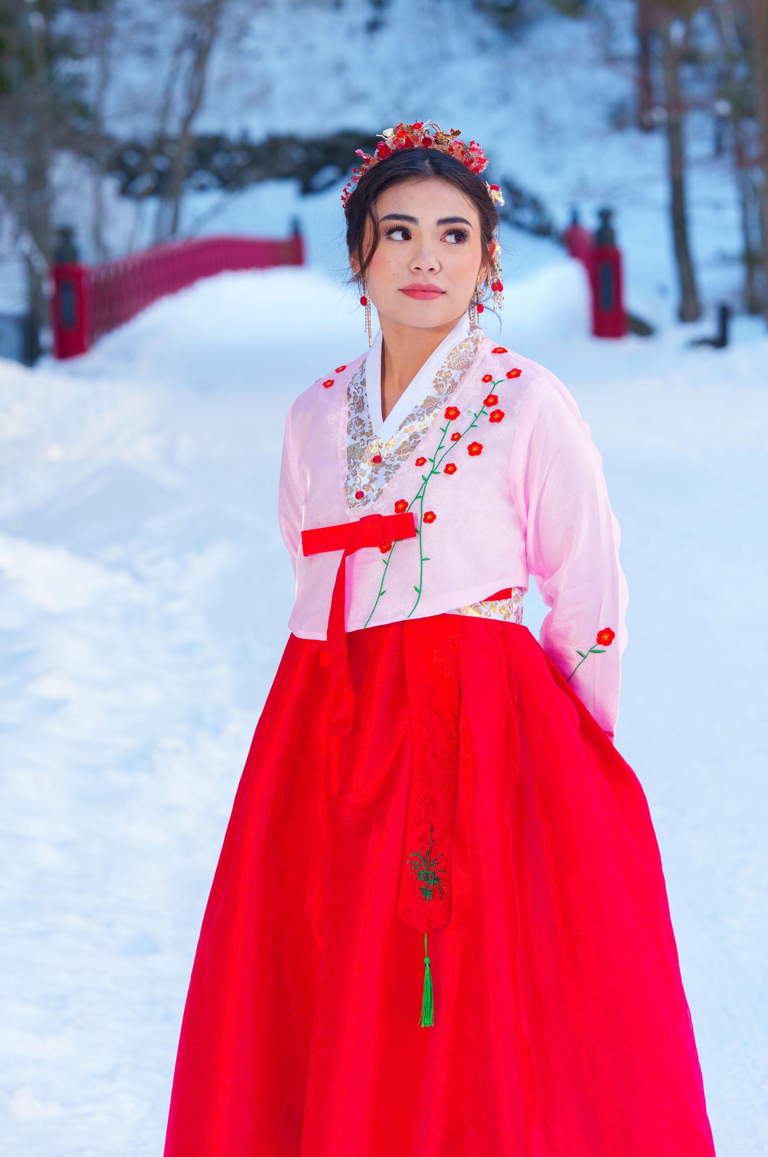 Woman in traditional Korean hanbok with red skirt and floral crown poses in snowy winter landscape.