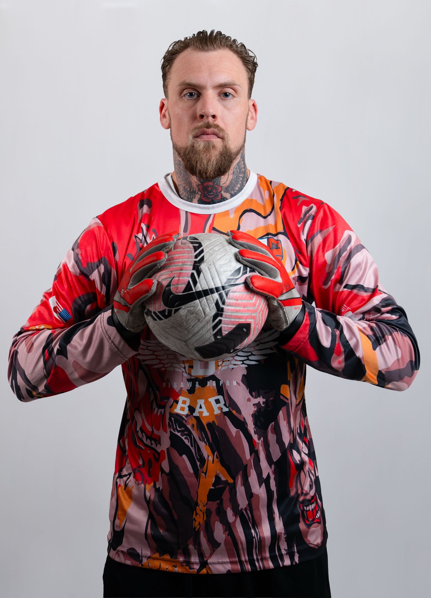 A tattooed goalkeeper in a vibrant red patterned long-sleeve jersey holding a soccer ball.