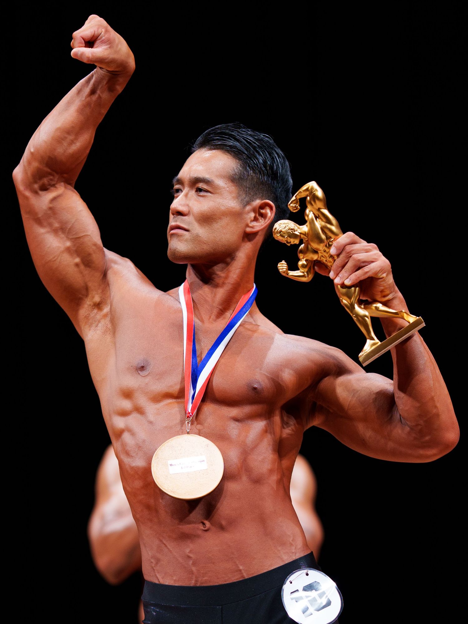 Muscular male bodybuilder celebrates victory holding gold trophy statue while wearing competition medals on dark stage.