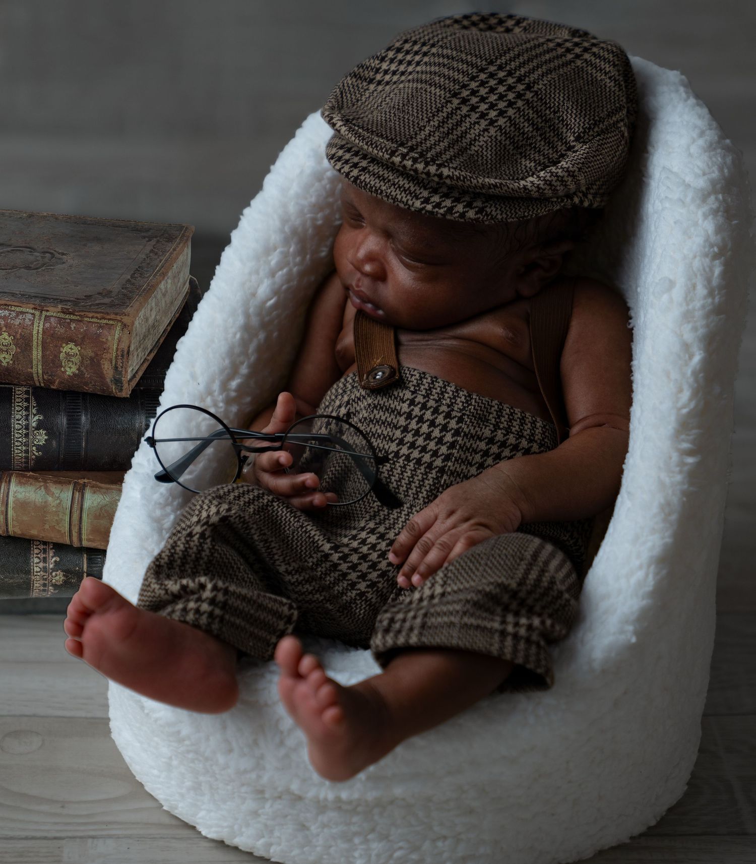 Adorable newborn baby dressed in houndstooth outfit and cap sleeps in cozy white chair with books.