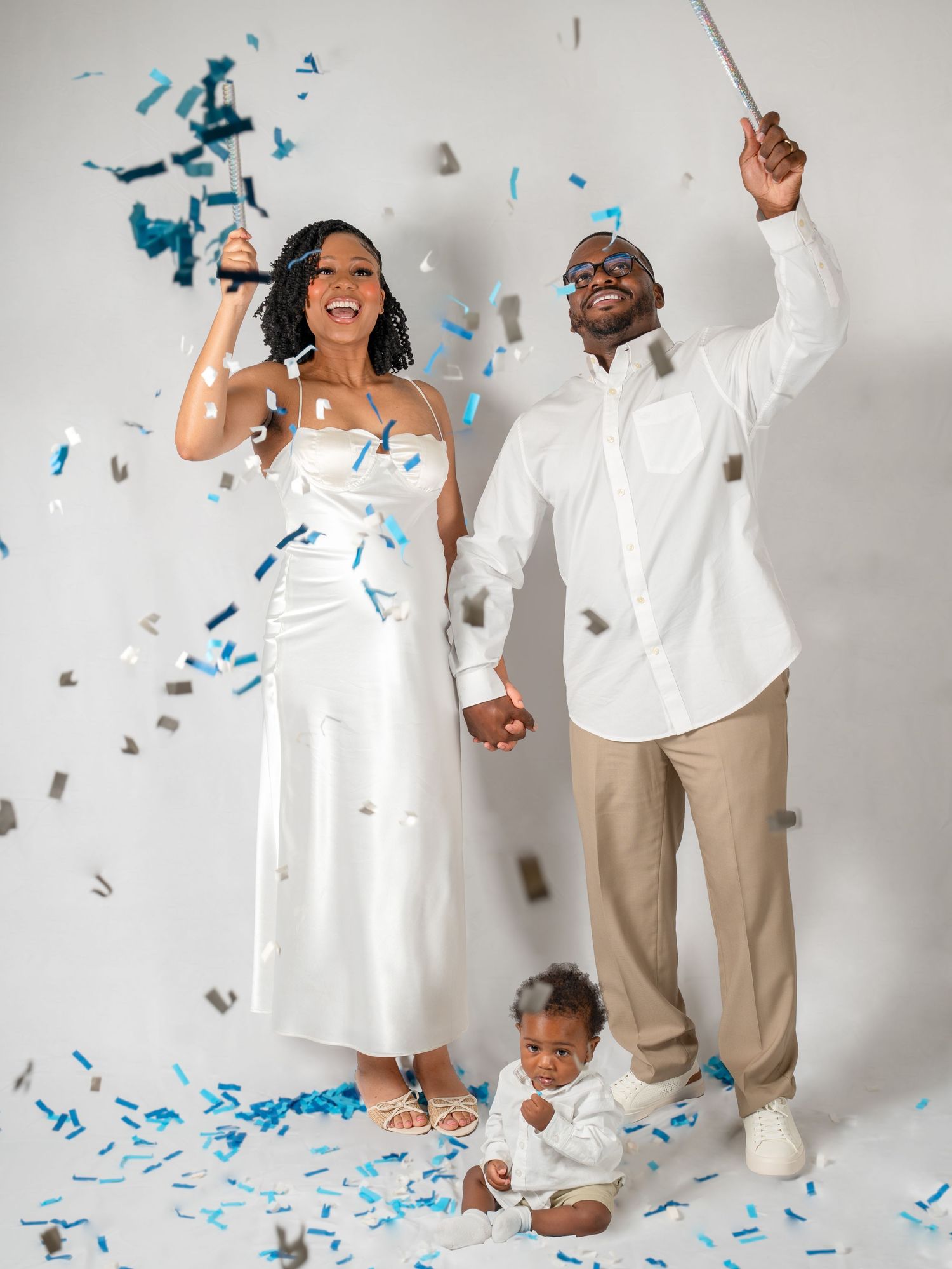 Happy couple with baby celebrates gender reveal with blue and silver confetti in white studio.