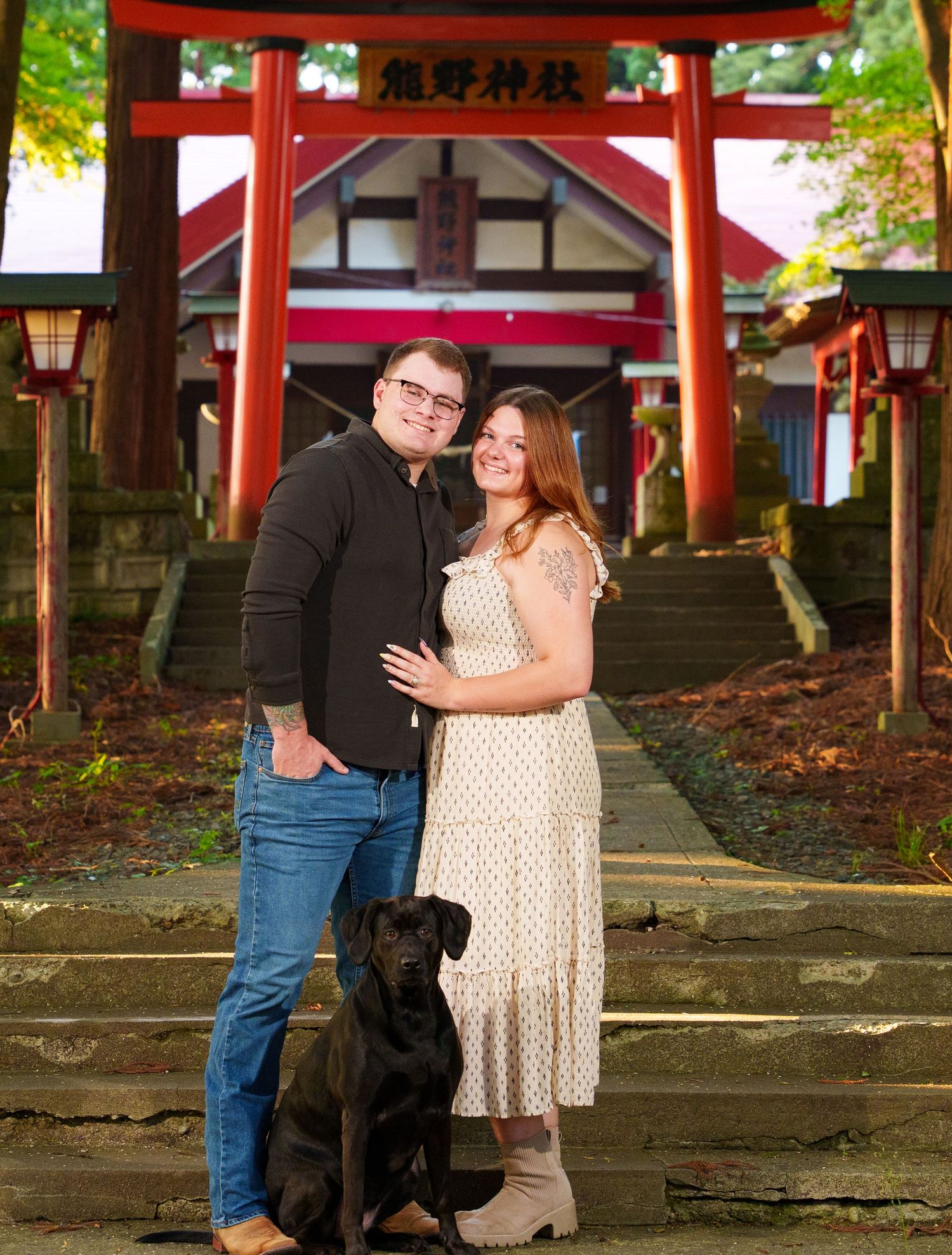 Couple with black Labrador poses in front of red Japanese torii gate shrine entrance at dusk.