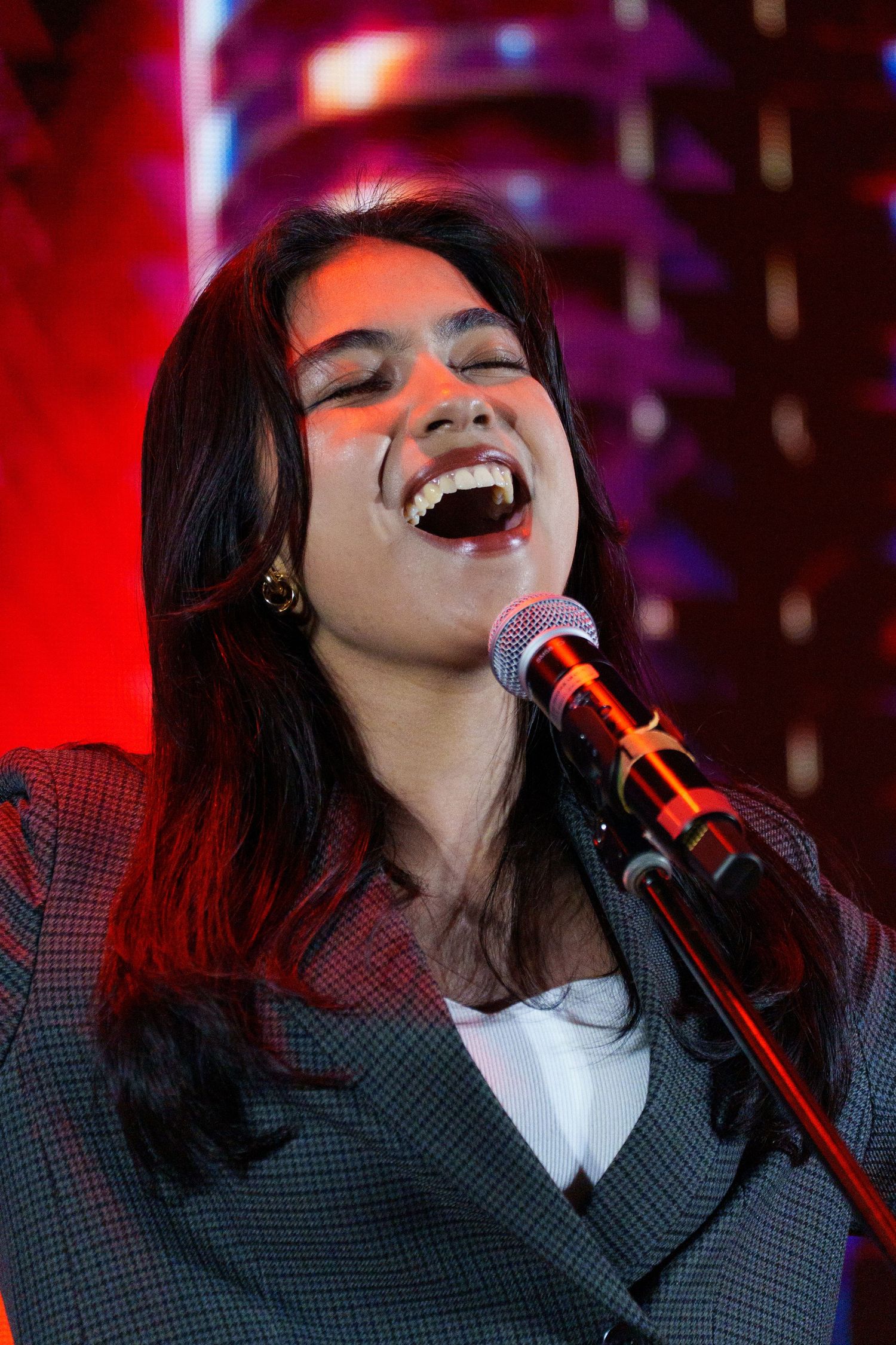 Young woman with dark hair performs passionately at microphone under dramatic red stage lighting at live concert.
