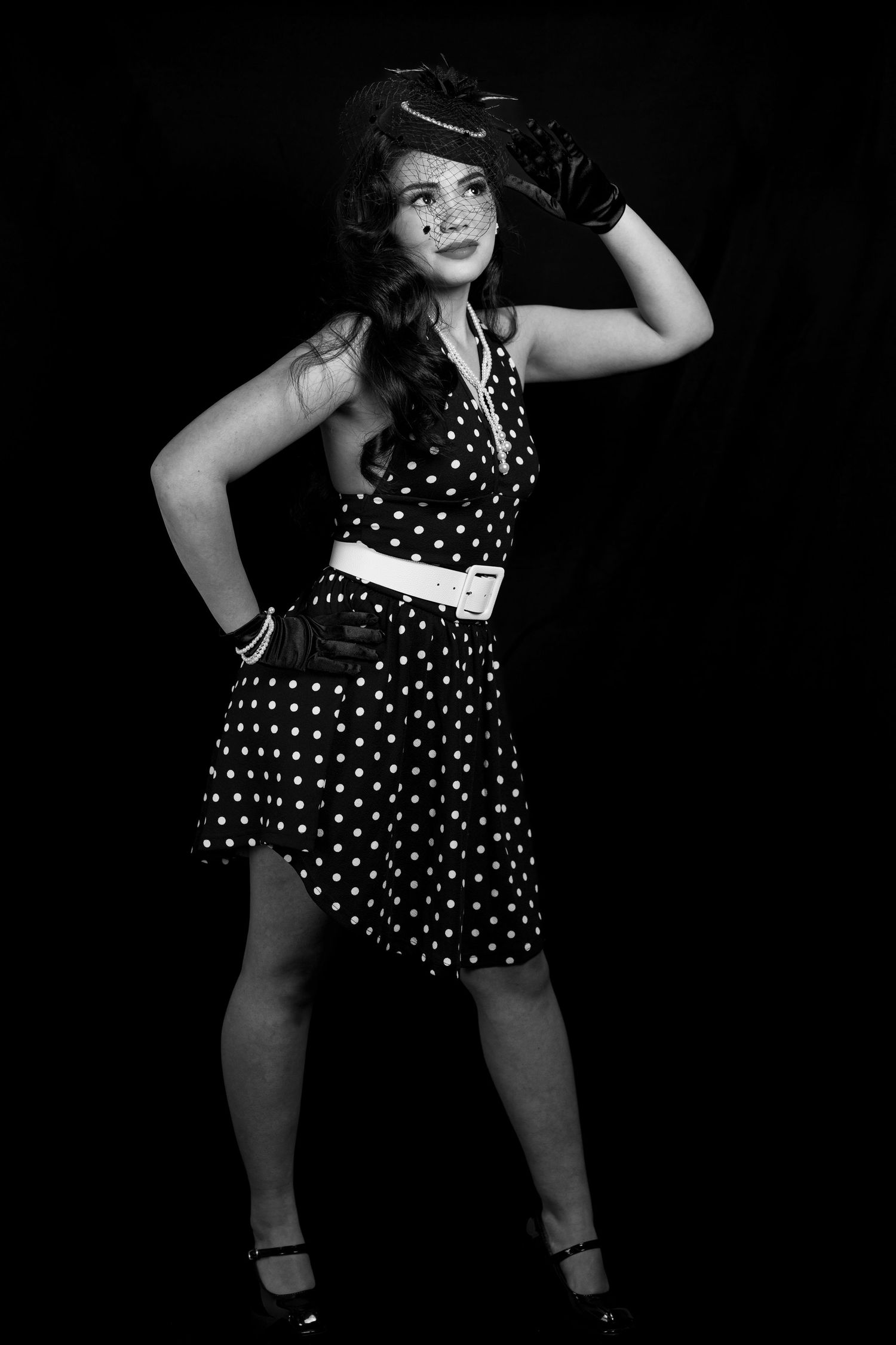 Vintage pin-up woman in polka dot dress and hat poses confidently in black and white studio photo.