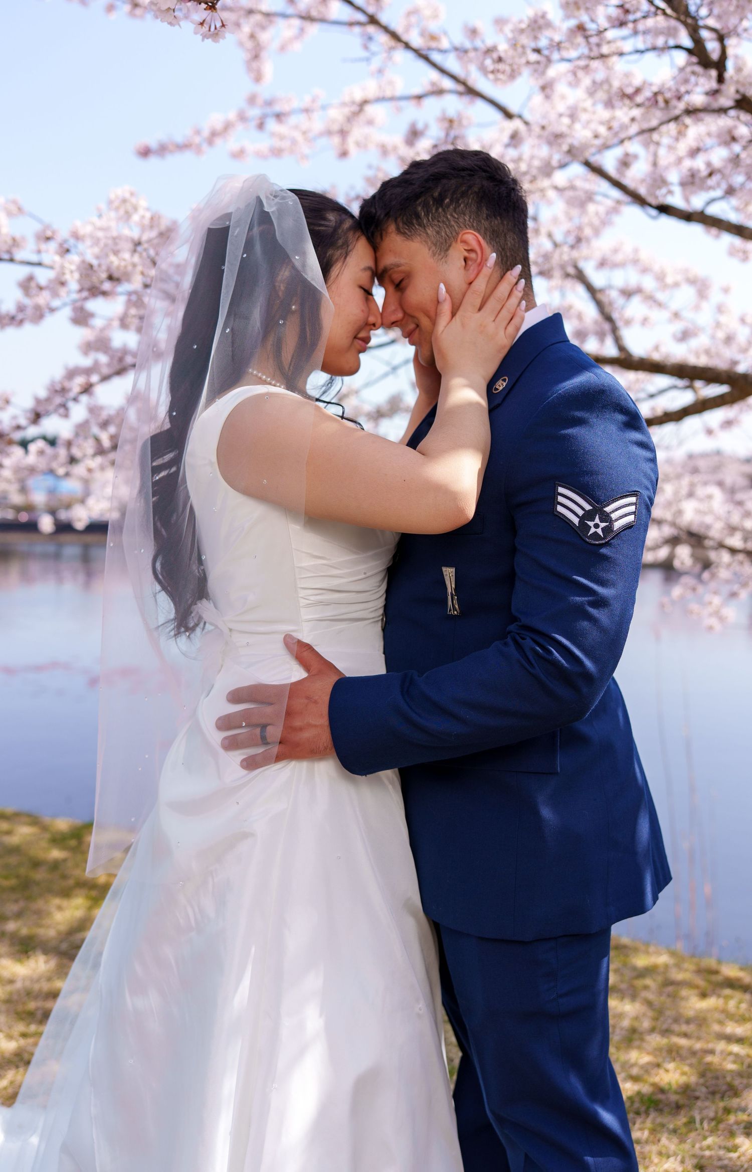 Bride and groom embracing tenderly under blooming cherry blossom trees in spring, groom in Air Force uniform.