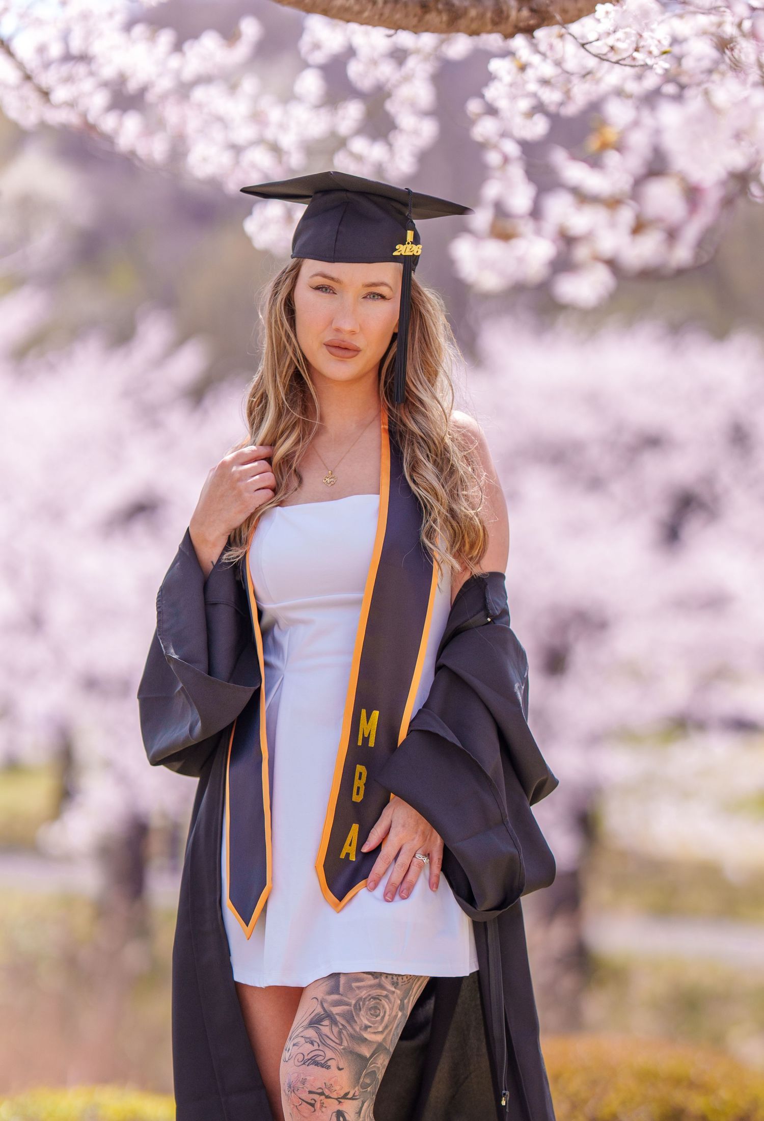 MBA graduate in cap and gown with tattoos poses under blooming cherry blossom trees in spring.