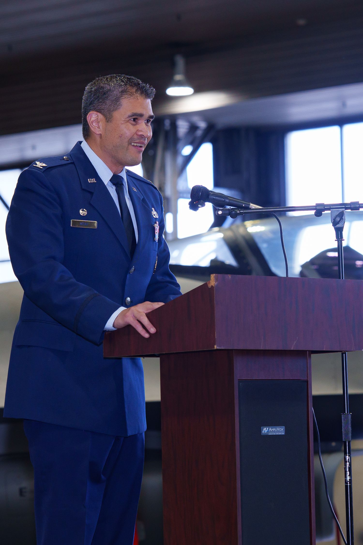 Military officer in blue Air Force uniform delivers speech at podium in aviation hangar setting.
