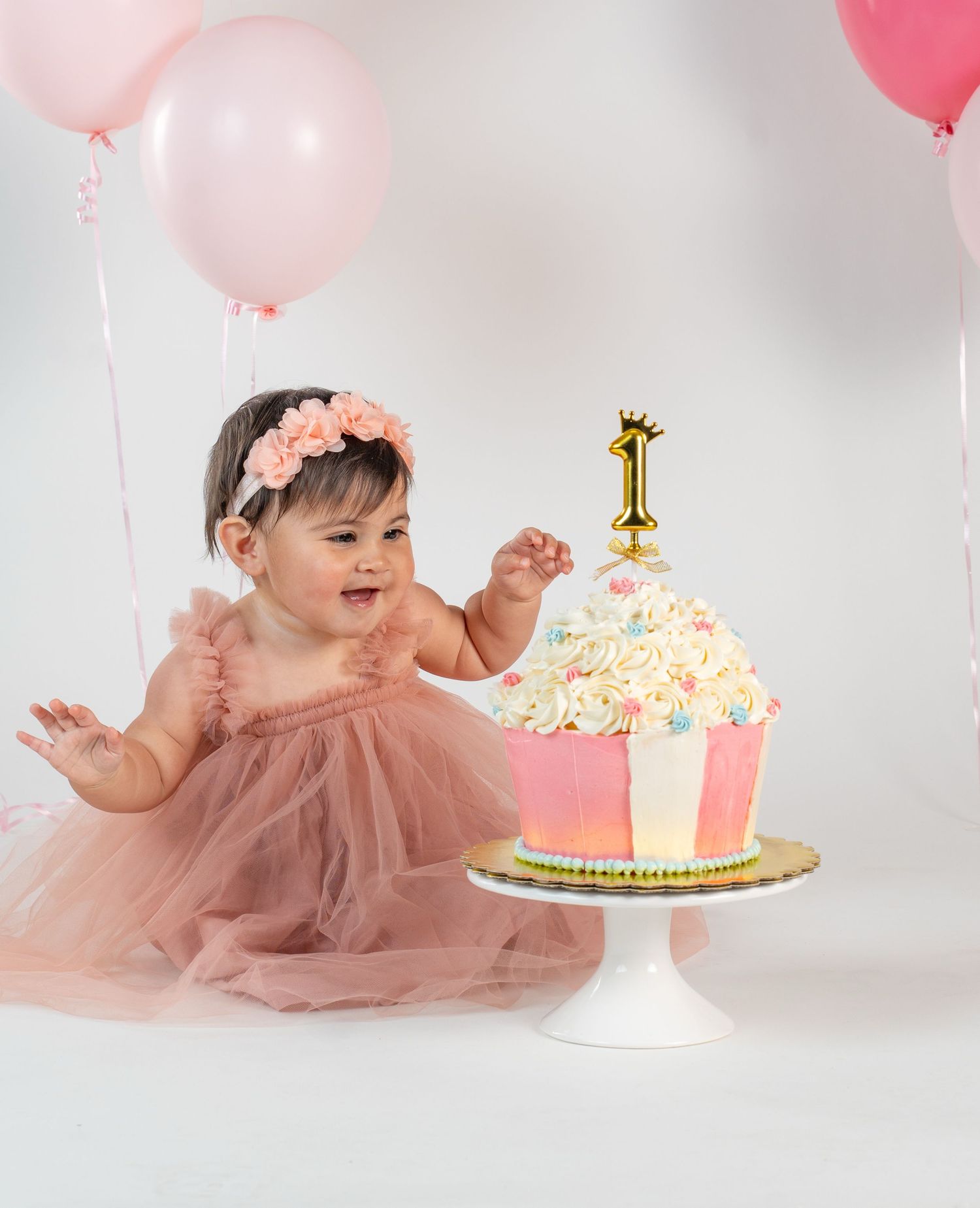 Baby girl in pink tutu celebrates first birthday with oversized cupcake and balloons.