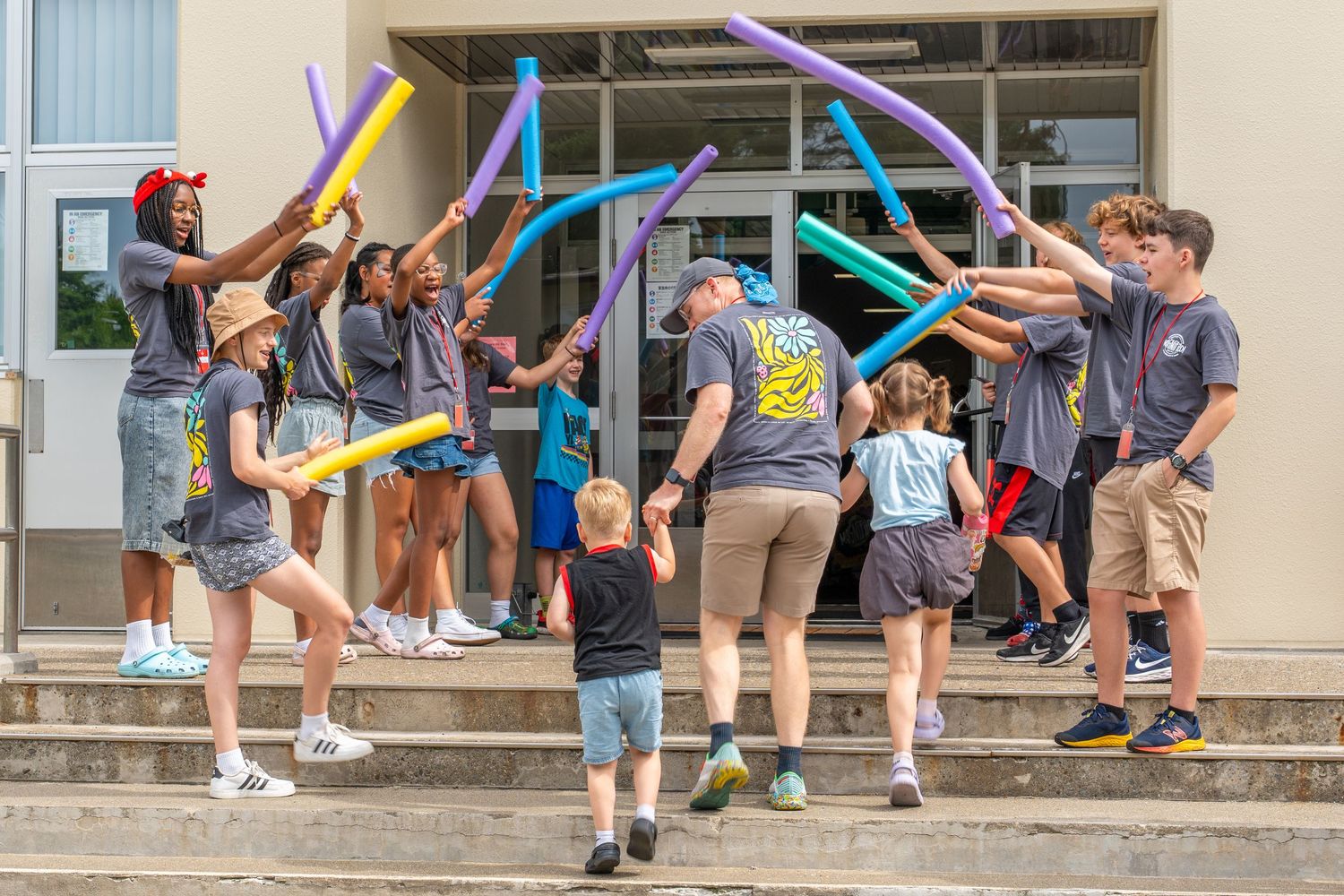 Group of teens with colorful balloon sticks welcoming children on outdoor building steps.