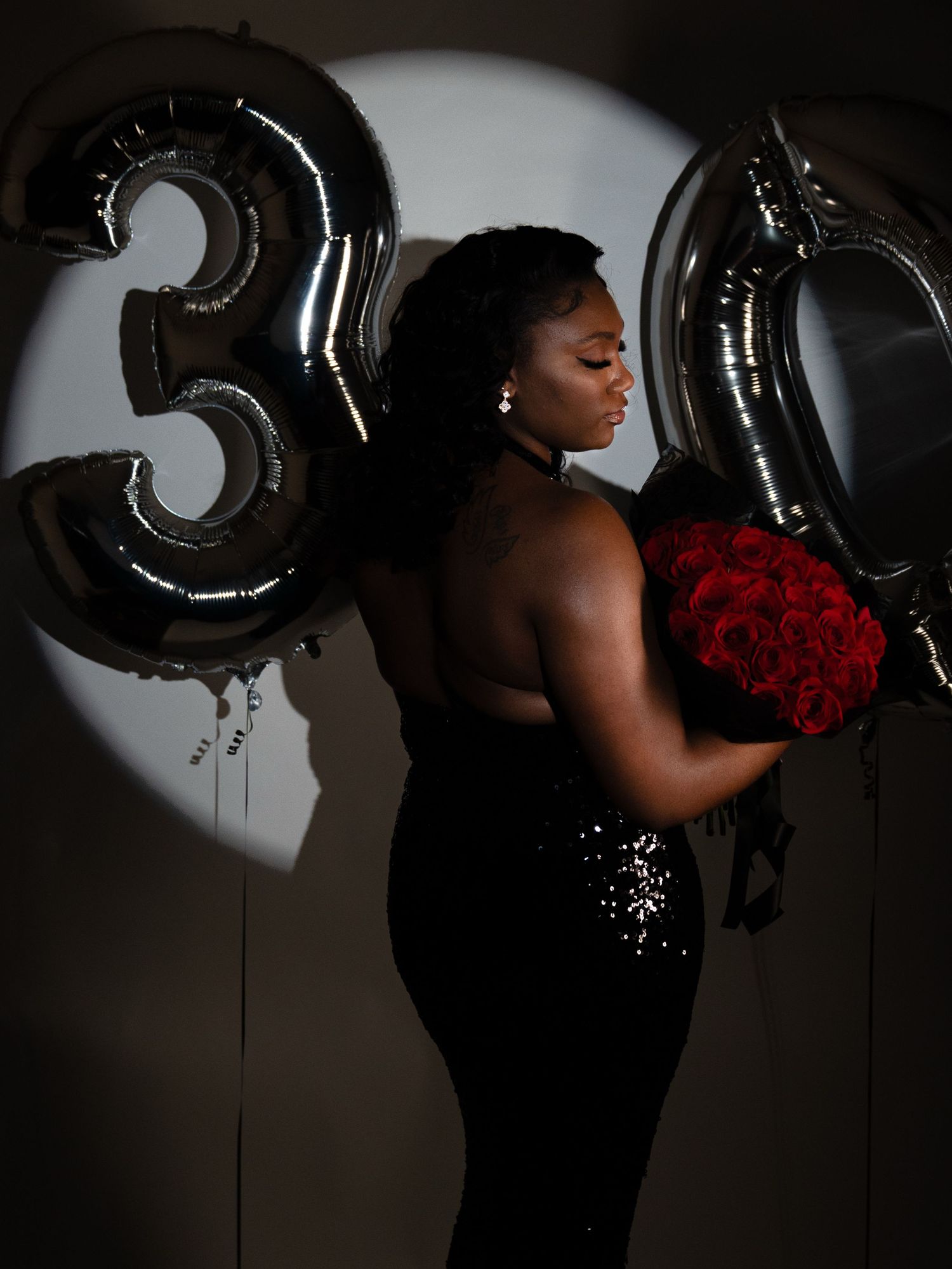 Woman in black sequin dress holds red roses beside silver 30th birthday balloons.