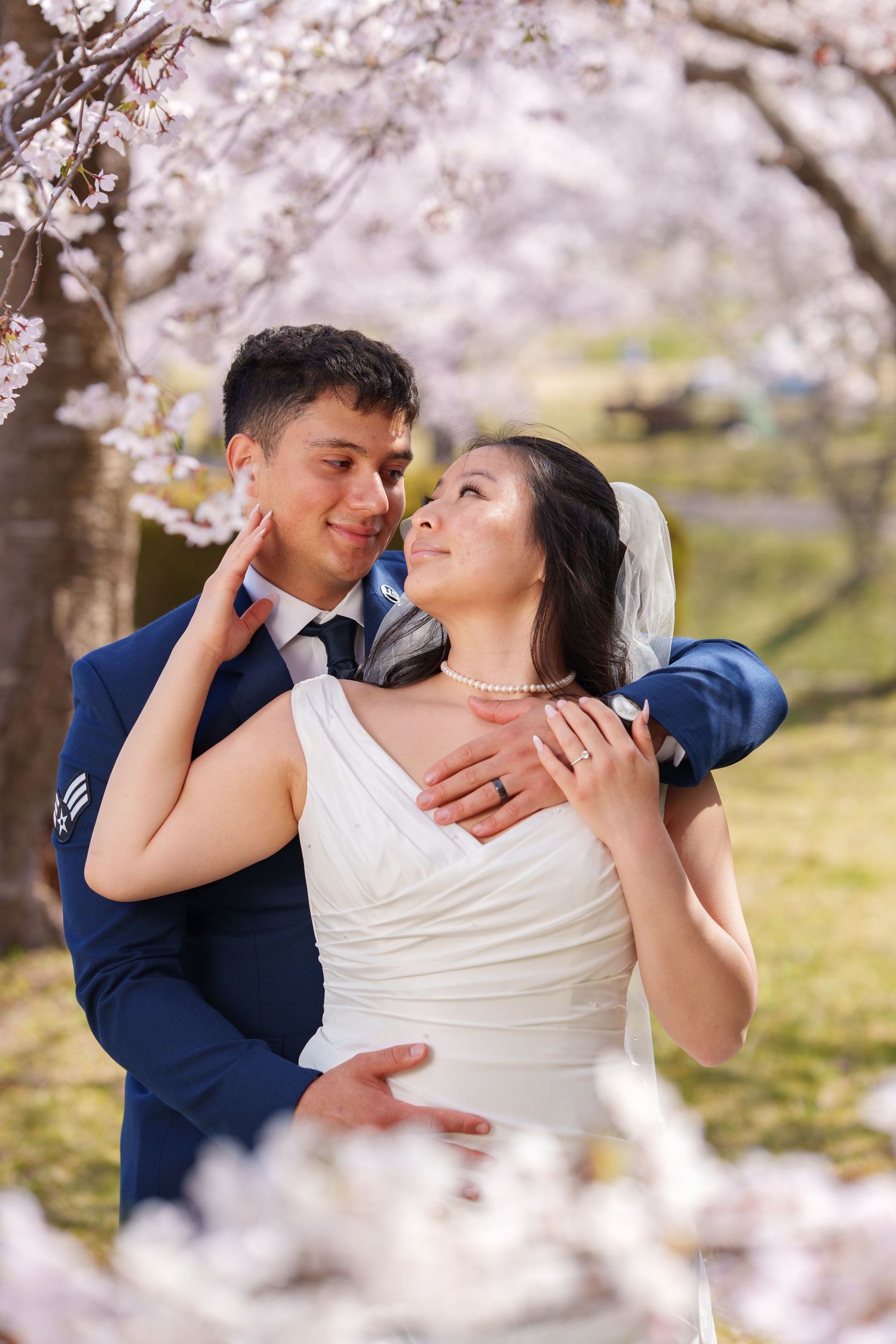 Groom in blue suit embracing bride in white dress with pearl necklace under pink cherry blossom trees.