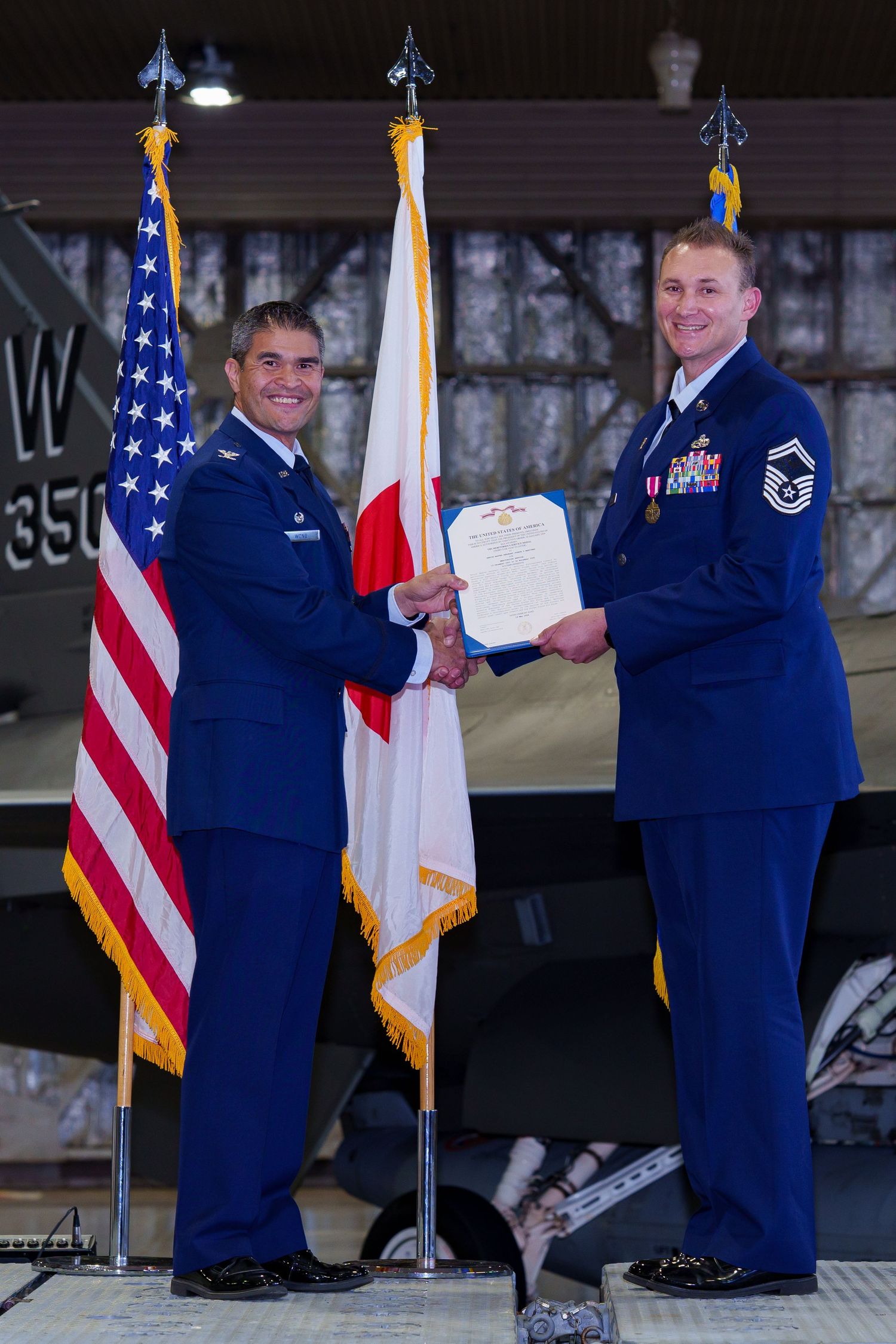 Two US Air Force officers in blue uniforms exchange a military certificate during an official ceremony with American and Japanese flags.