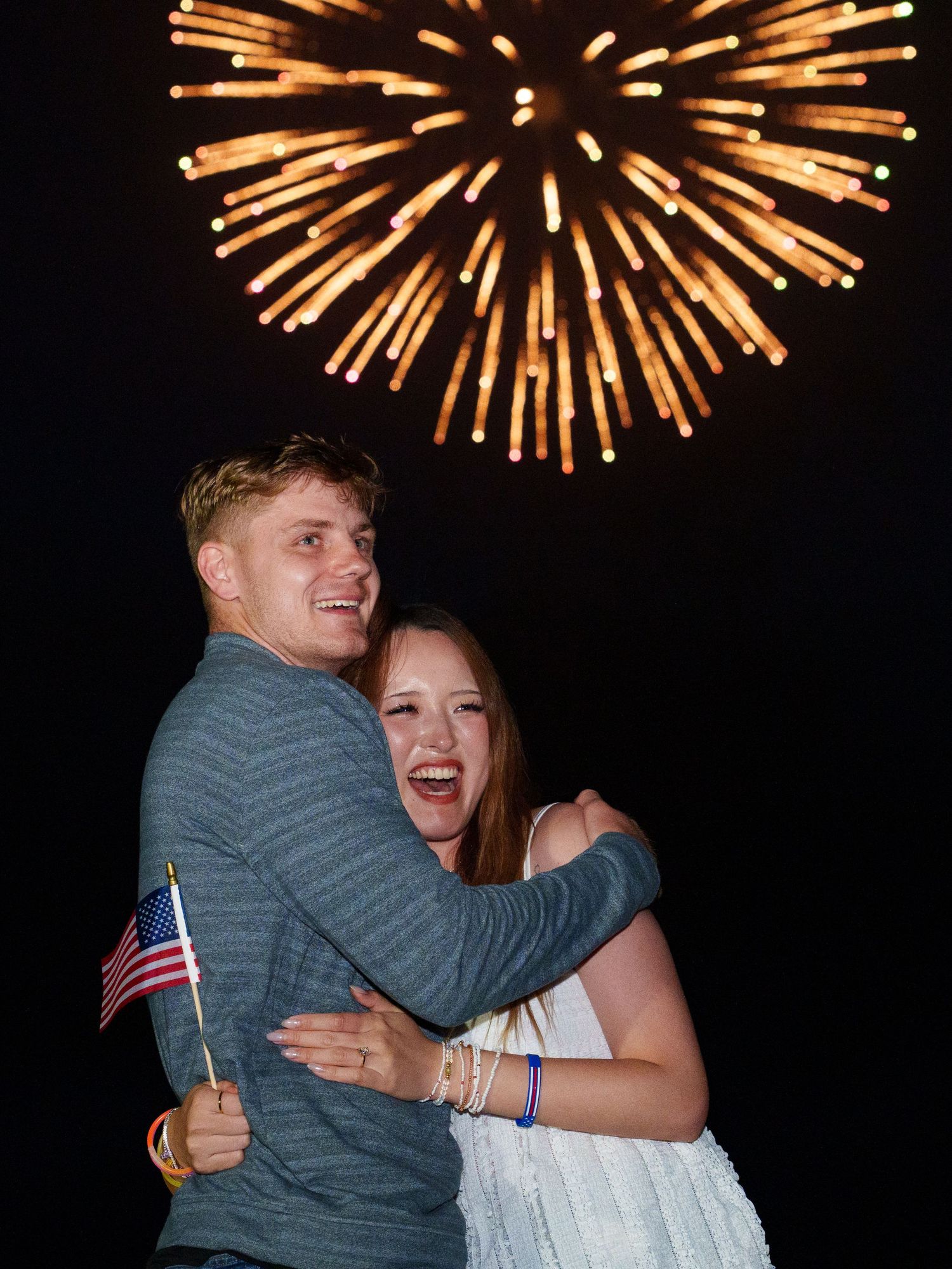 Happy couple embracing and laughing while holding American flag under golden fireworks in night sky.