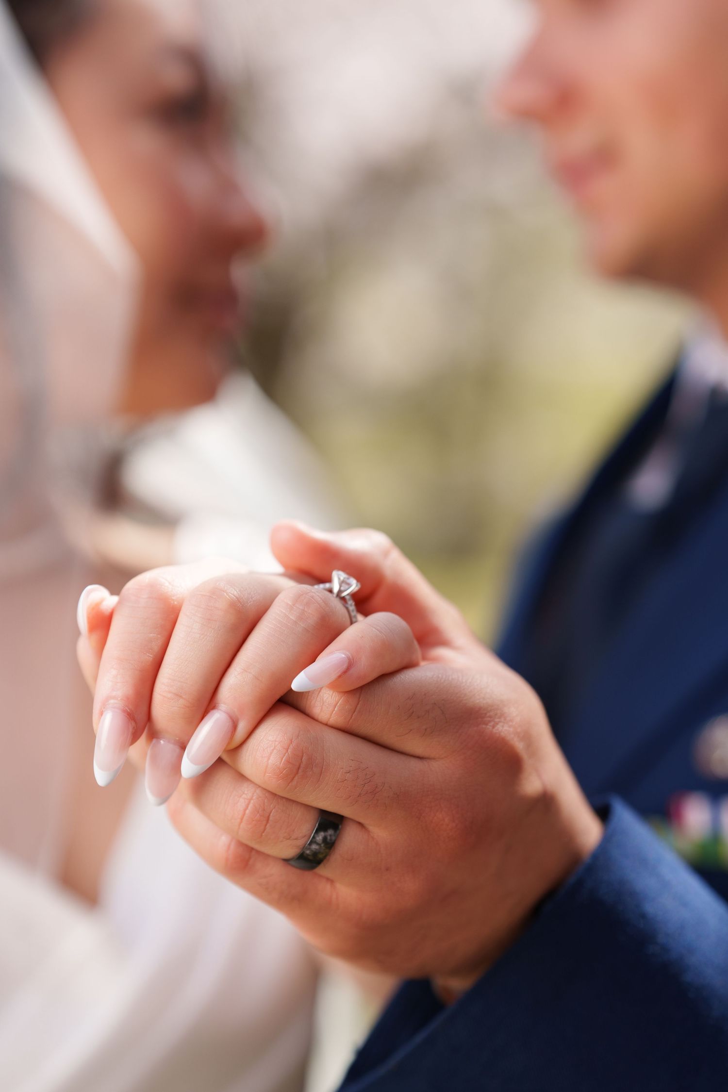 Close-up of newlyweds holding hands showing diamond engagement ring and black wedding band outdoors.