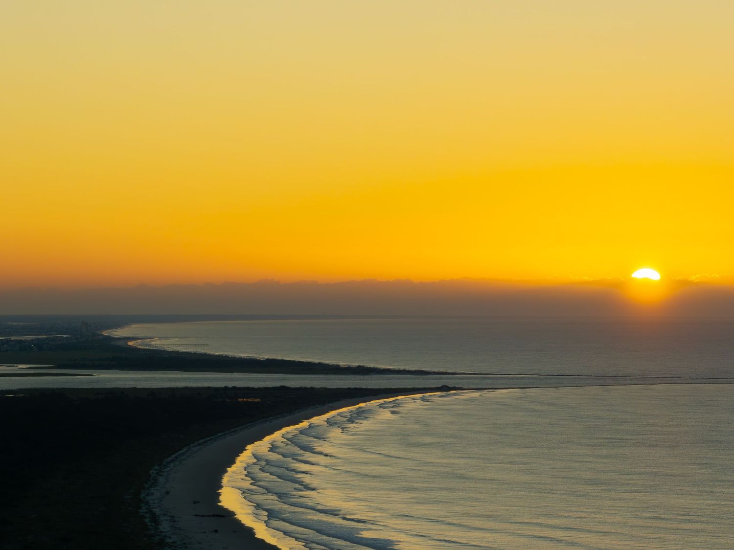 Stunning aerial sunset view over a curved coastline with golden sky reflecting on calm ocean waters.