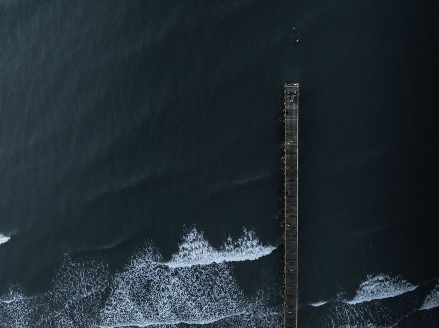 Aerial drone view of a long wooden pier extending into dark ocean waters with crashing waves.