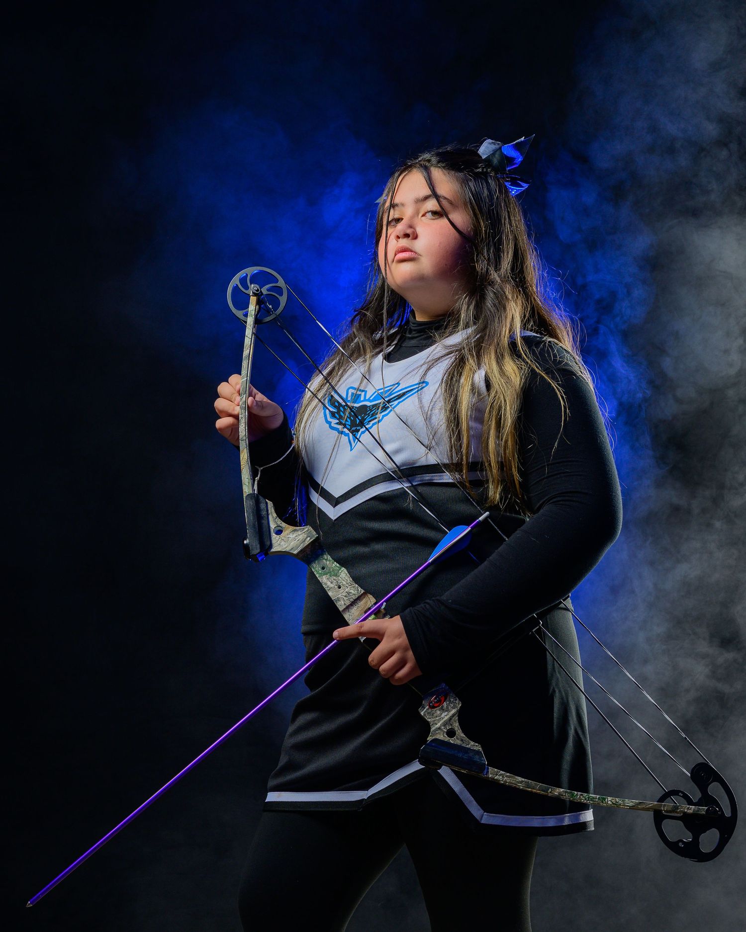 Girl in cheerleader uniform poses dramatically with compound bow amid blue smoke on dark background.