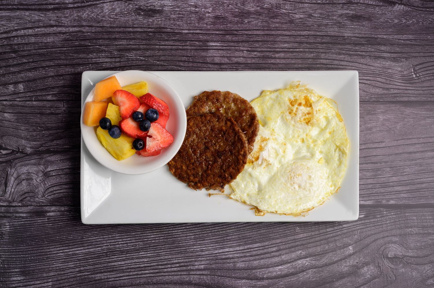 Breakfast plate with sunny-side-up eggs, hash brown patty, and fresh fruit bowl with berries and melon on white plate.