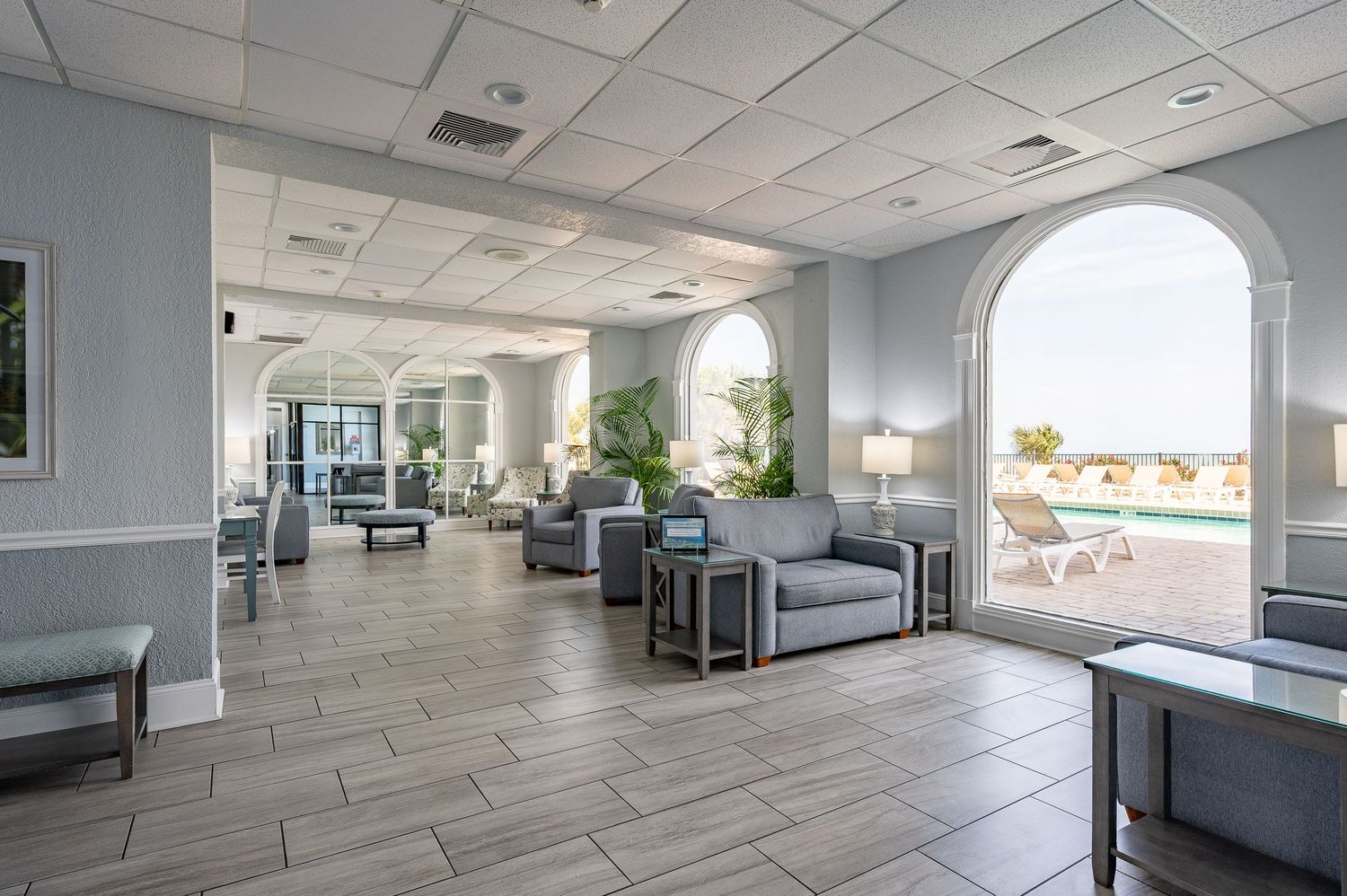 Spacious lobby lounge with gray seating, arched windows, and tile flooring in a coastal building.