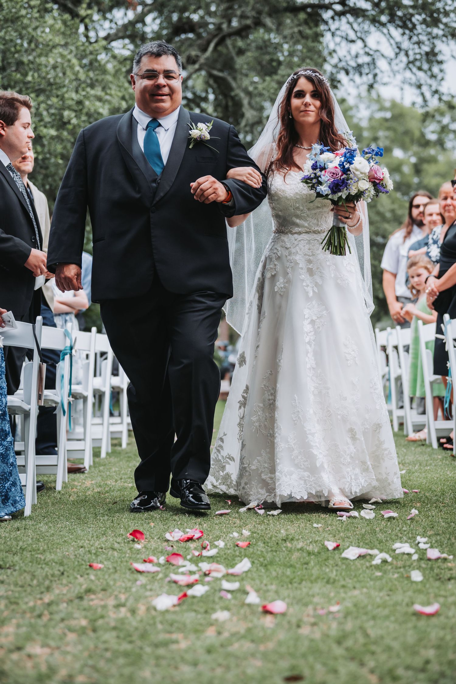 Bride walks down outdoor wedding aisle with escort, holding colorful bouquet, rose petals on grass.
