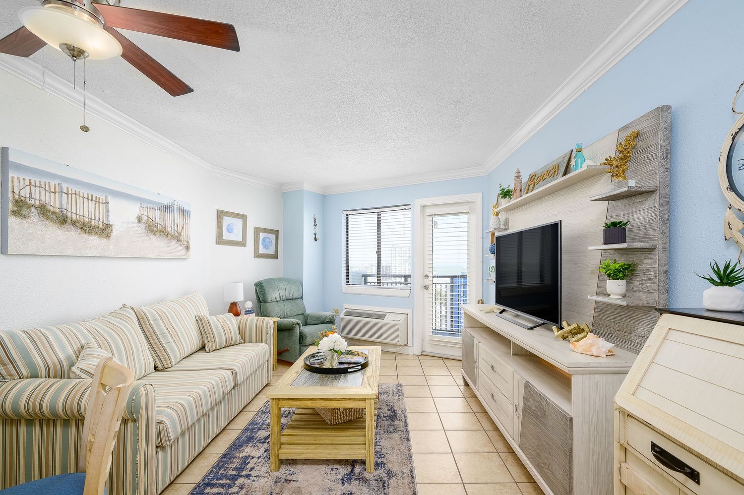 Bright beachside living room with striped sofa, TV unit, ceiling fan, and ocean view balcony.