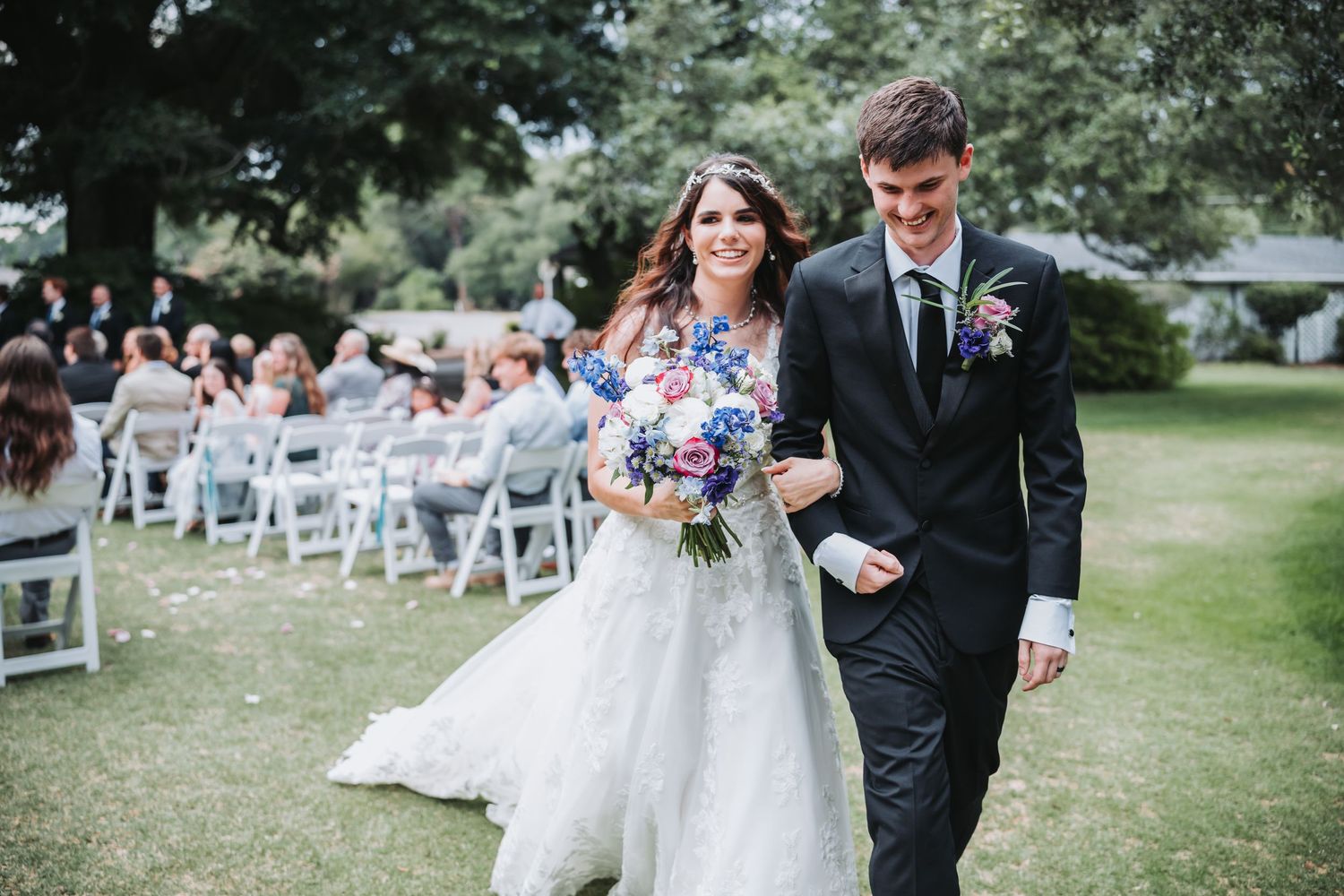 Newlyweds walk joyfully down outdoor wedding aisle after ceremony, bride holding colorful bouquet.