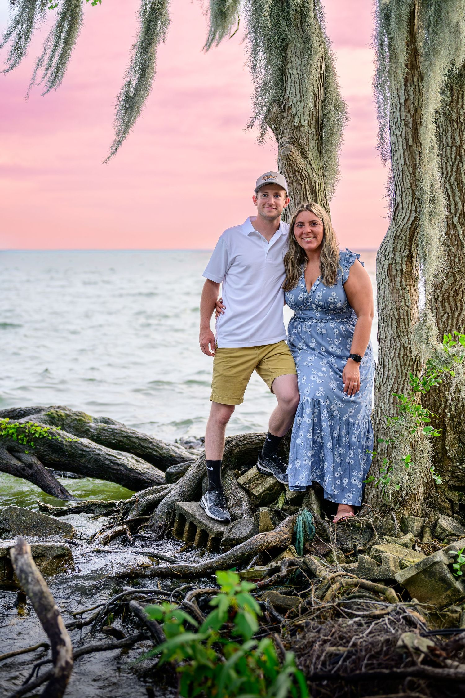 Smiling couple posing under Spanish moss tree by ocean at pink sunset.