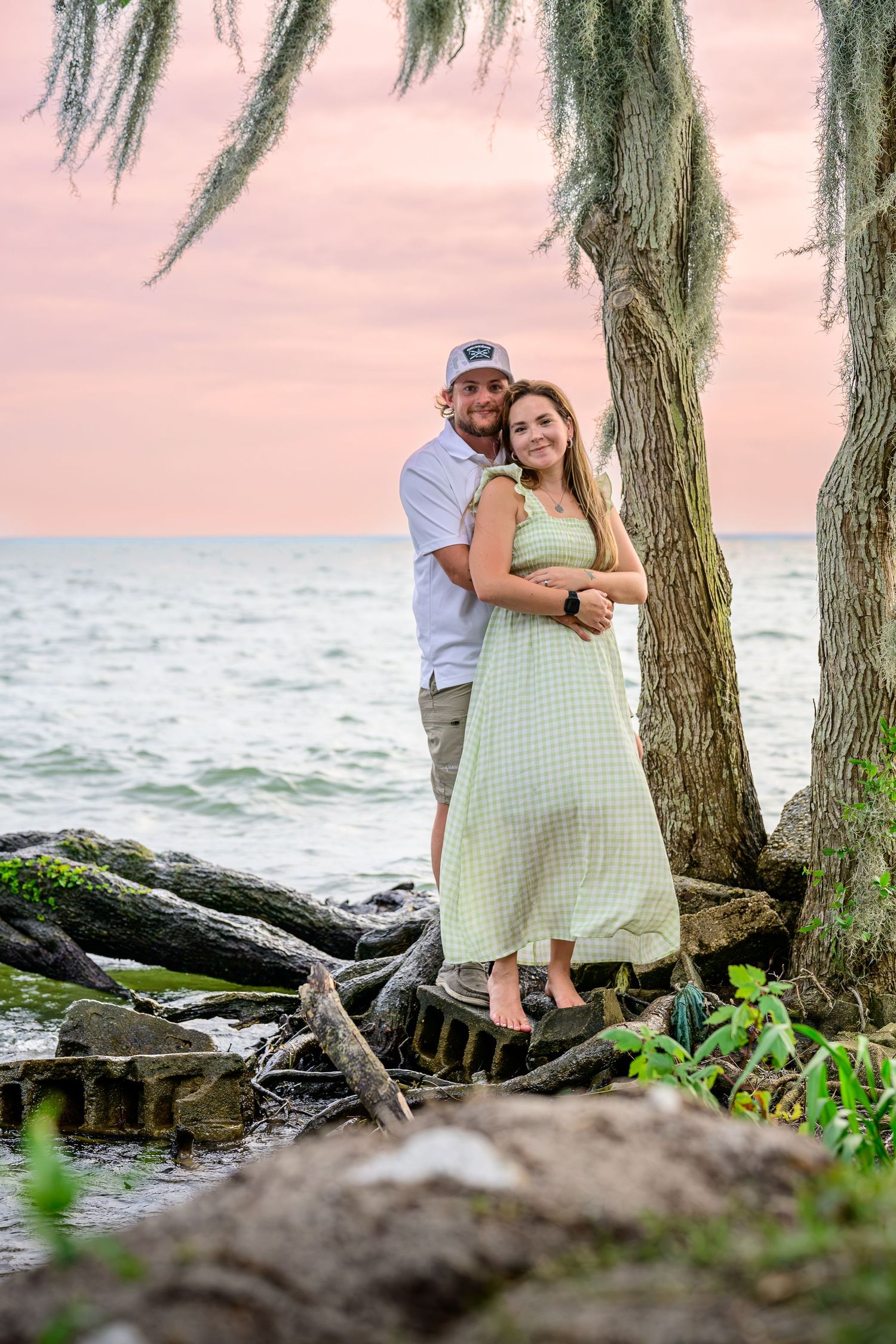 Couple embracing under Spanish moss tree at rocky shoreline during pink sunset.