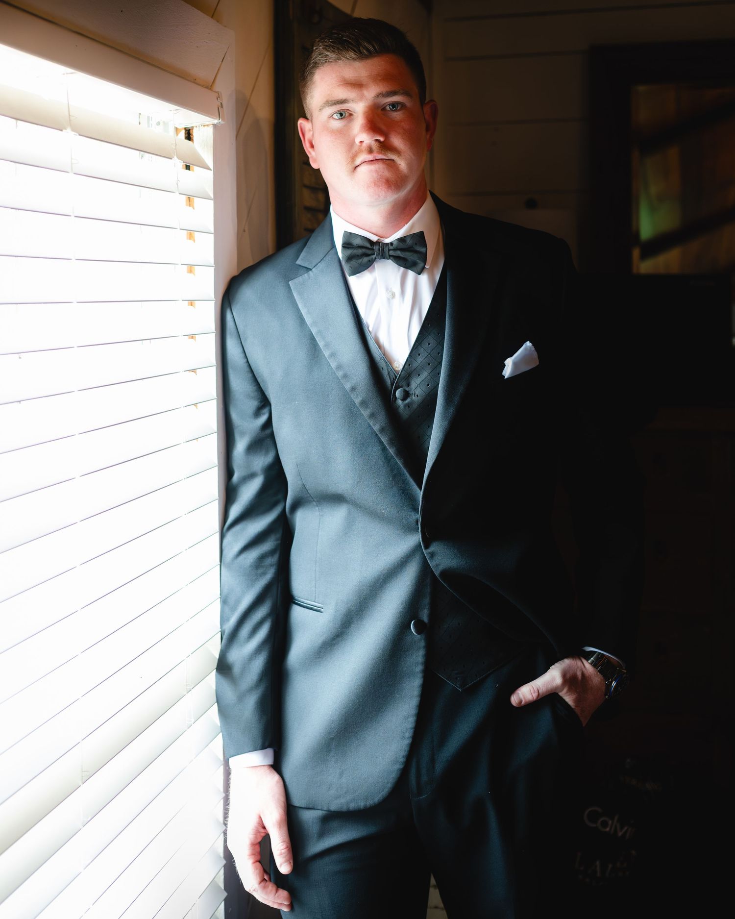 Handsome groom in dark tuxedo and bow tie posing by window in soft light.
