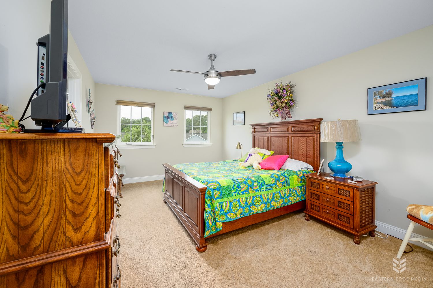 Cozy bedroom with tropical bedding, wooden furniture, ceiling fan, and carpet flooring near a bright window.