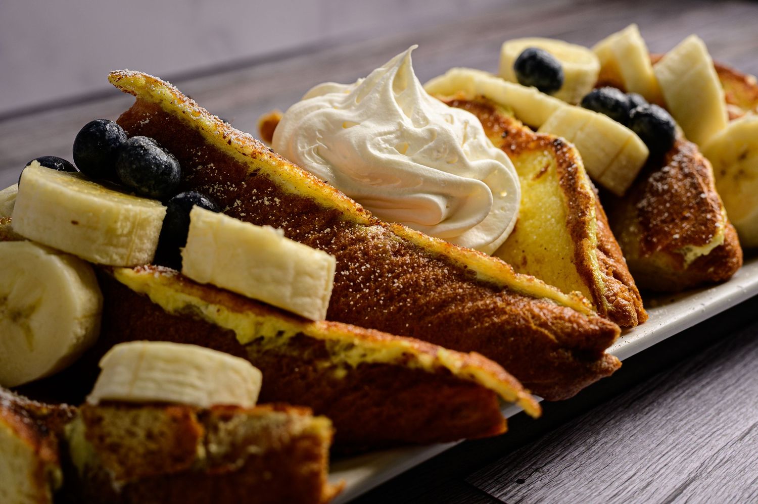 French toast slices topped with whipped cream, fresh banana slices, and blueberries on a white plate.