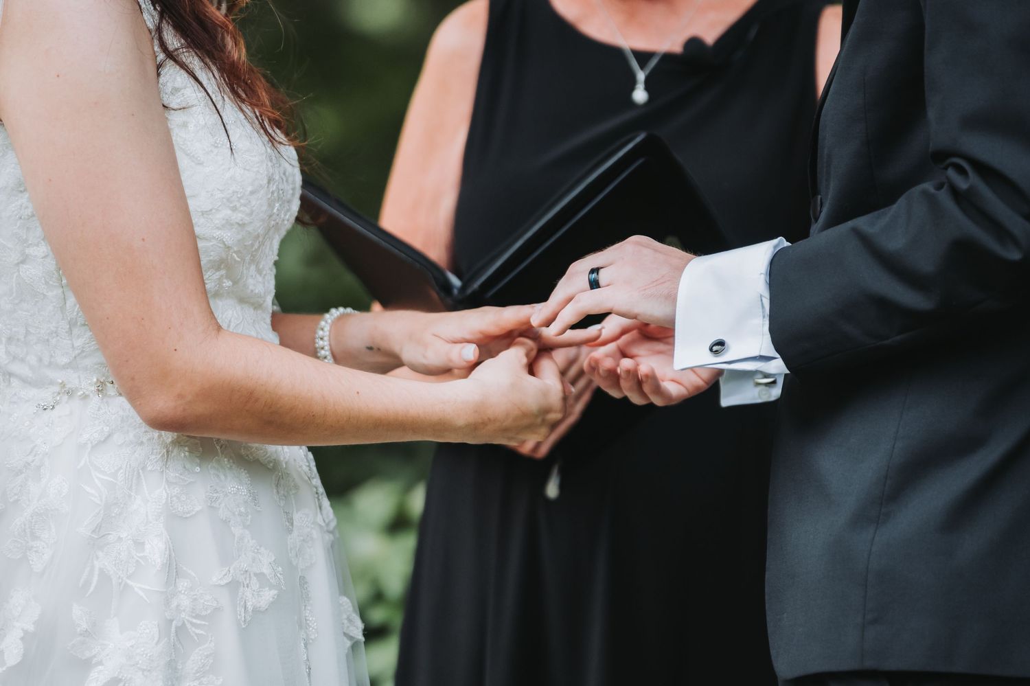 Officiant places wedding ring on bride's finger during intimate outdoor ceremony ring exchange.