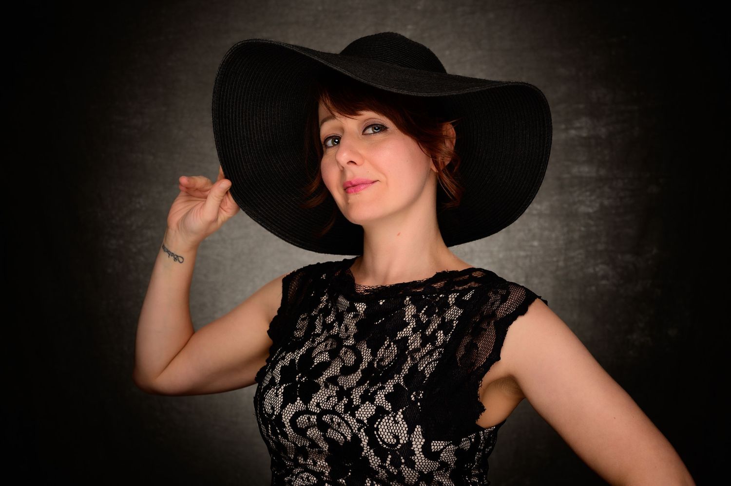 Elegant redhead woman in black lace dress poses with wide-brim black hat in studio.