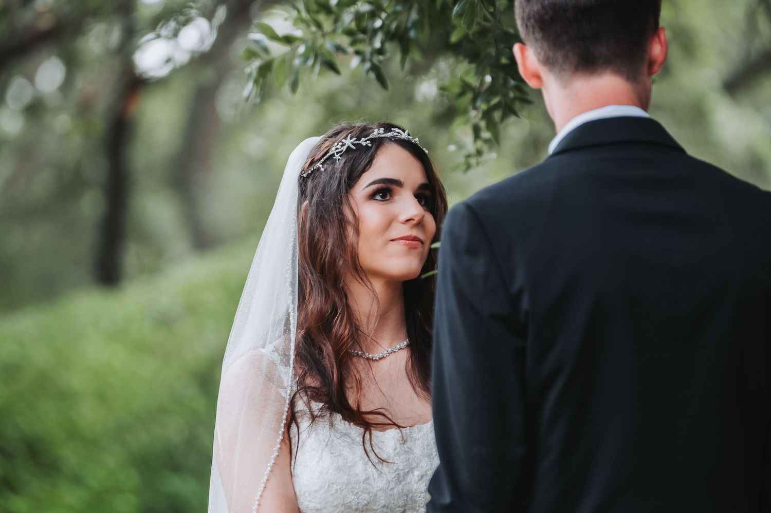 Bride in lace gown and veil gazes lovingly at groom during outdoor wedding ceremony among green trees.