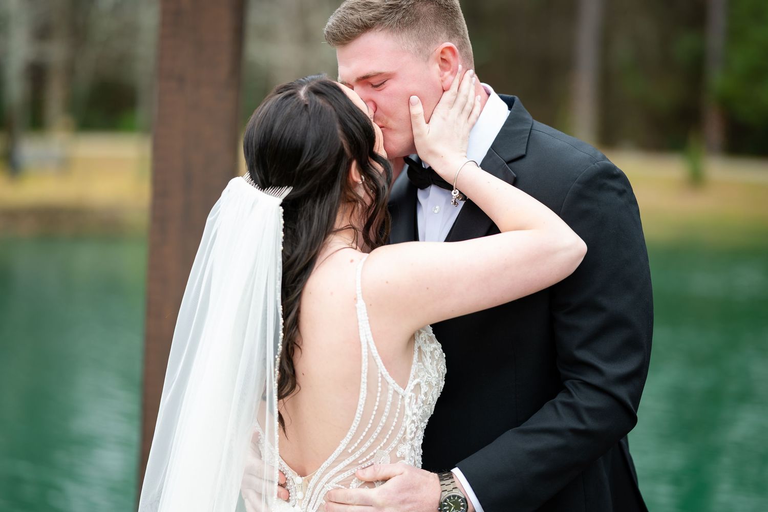 Bride and groom sharing first kiss outdoors near green lake after wedding ceremony.