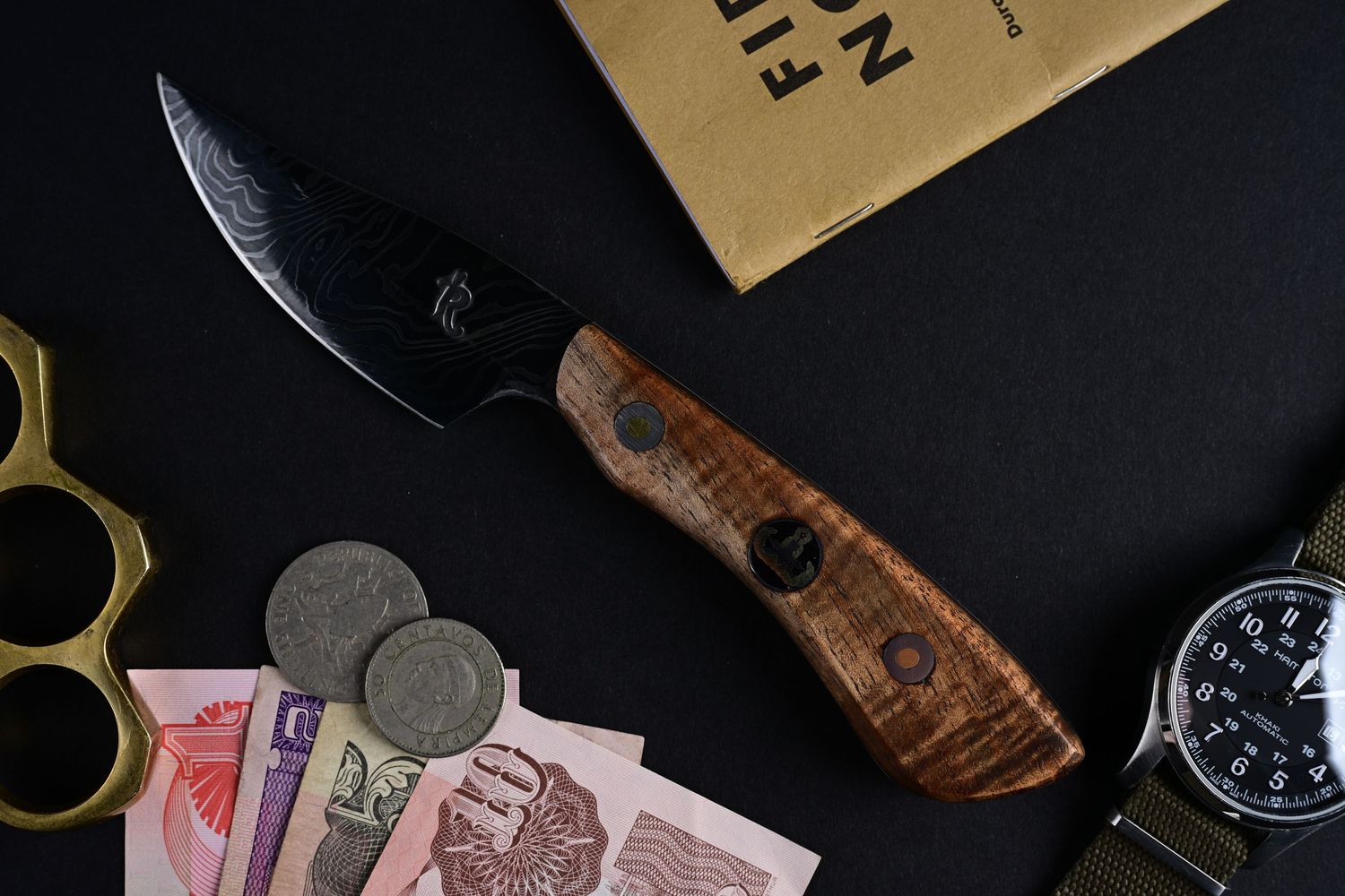 Handmade Damascus knife with wood handle displayed with brass knuckles, watch, coins, and currency notes.