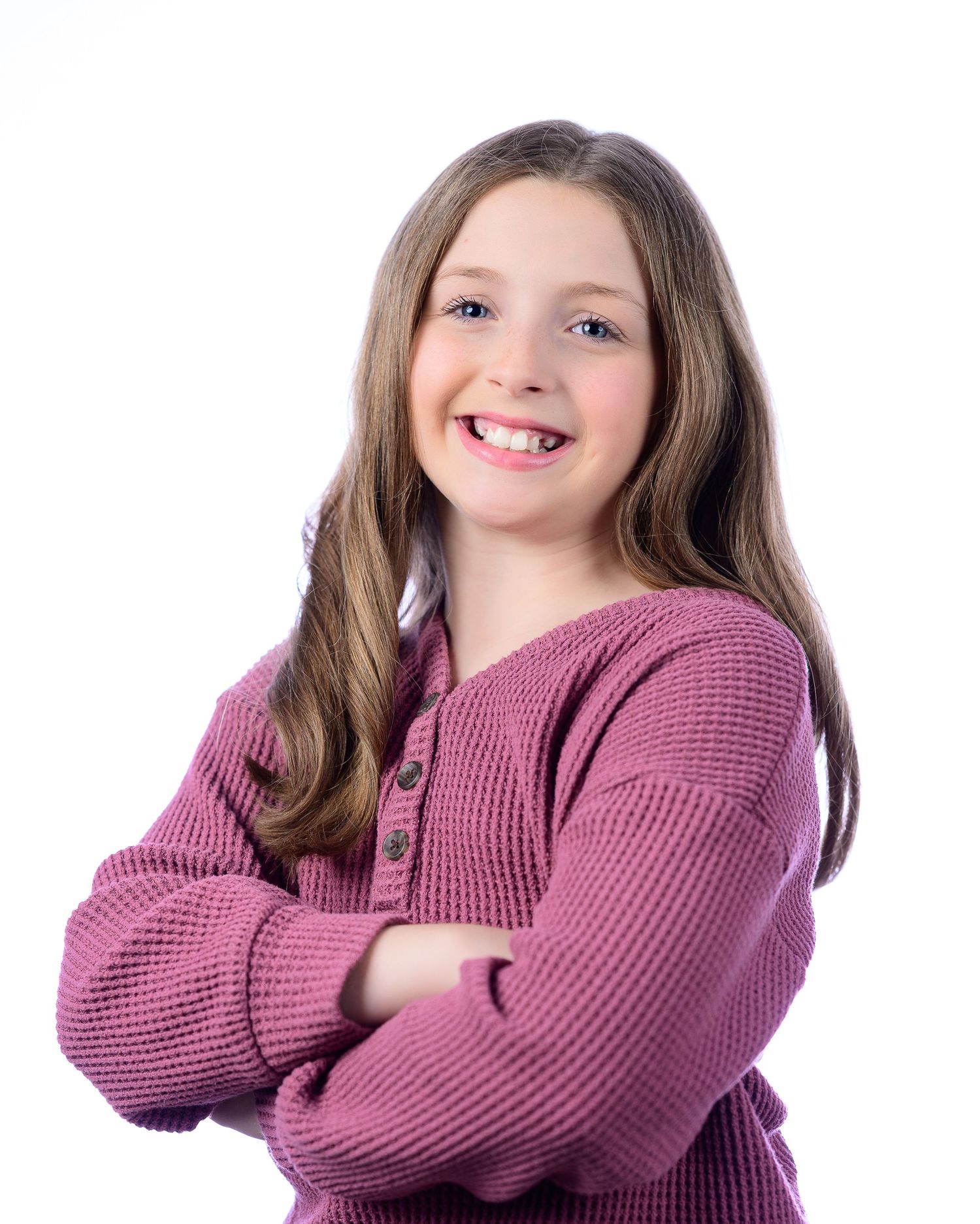 Smiling young girl with long brown hair poses with arms crossed wearing pink knit sweater.