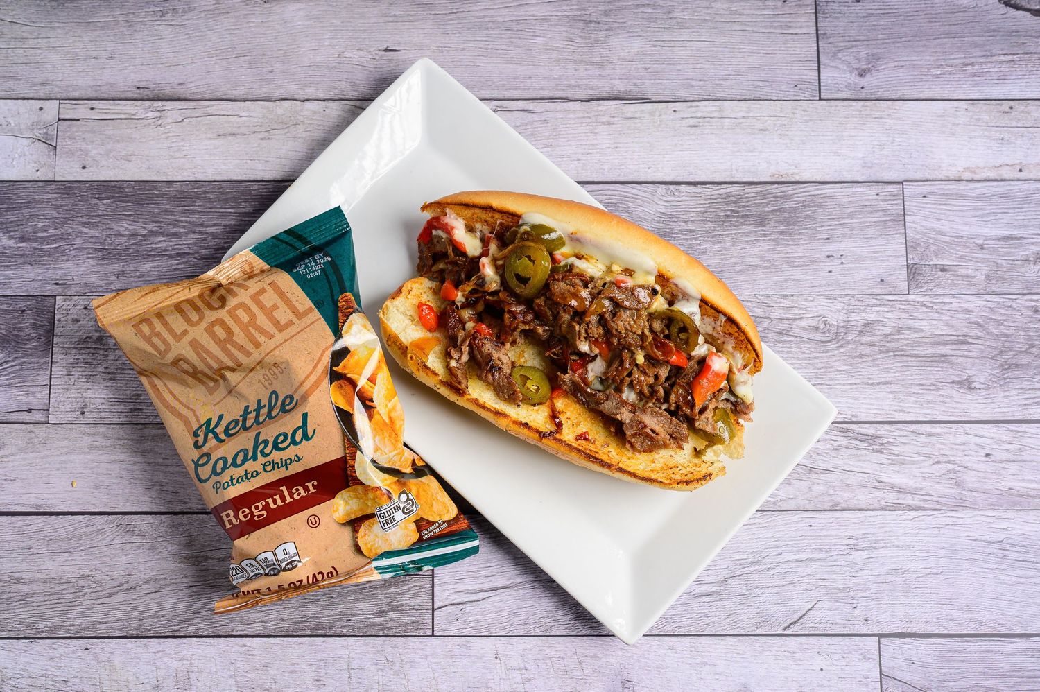 Philly cheesesteak sandwich with peppers and onions on a white plate beside kettle cooked potato chips bag.