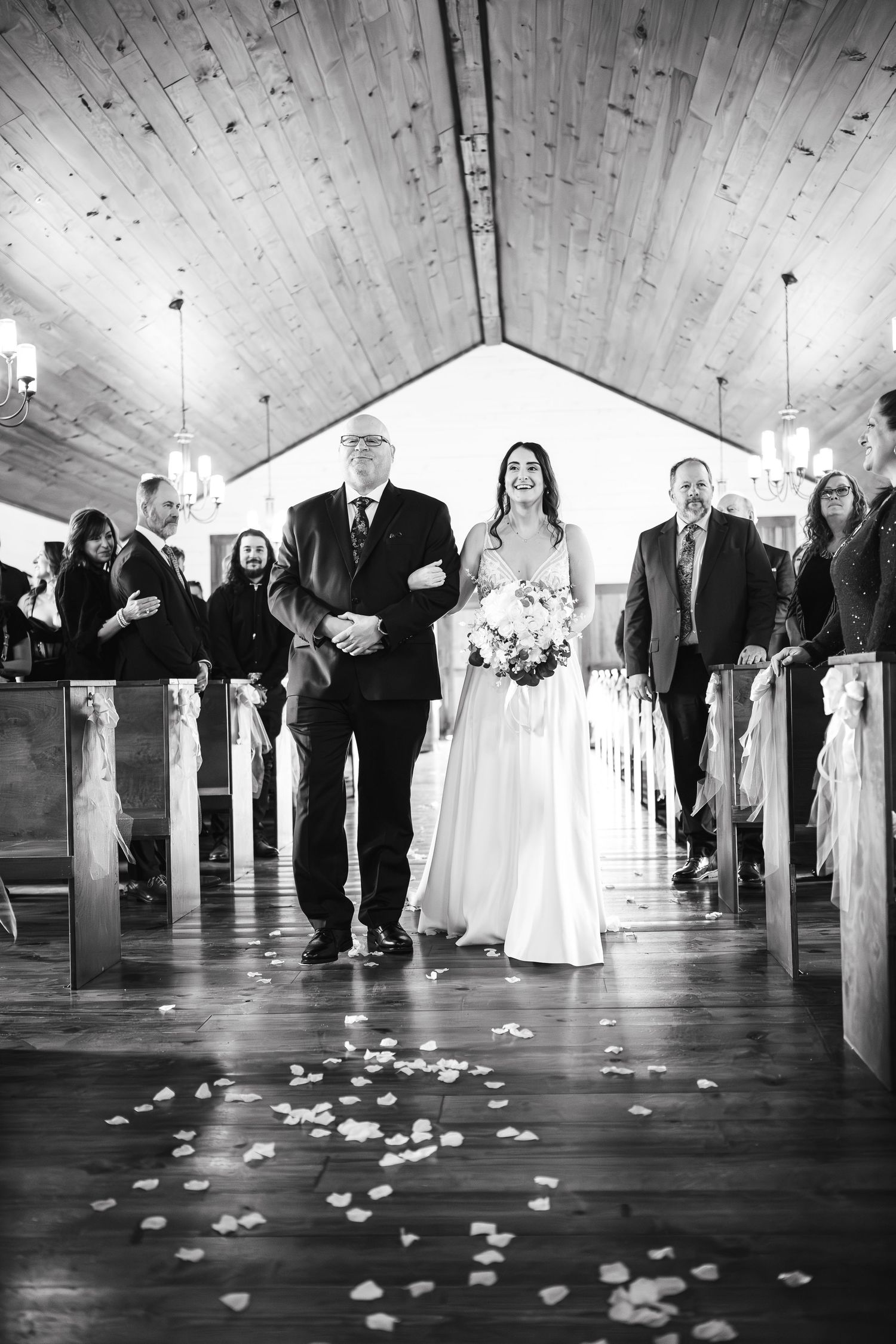 Black and white photo of bride walking down aisle with escort in rustic chapel.