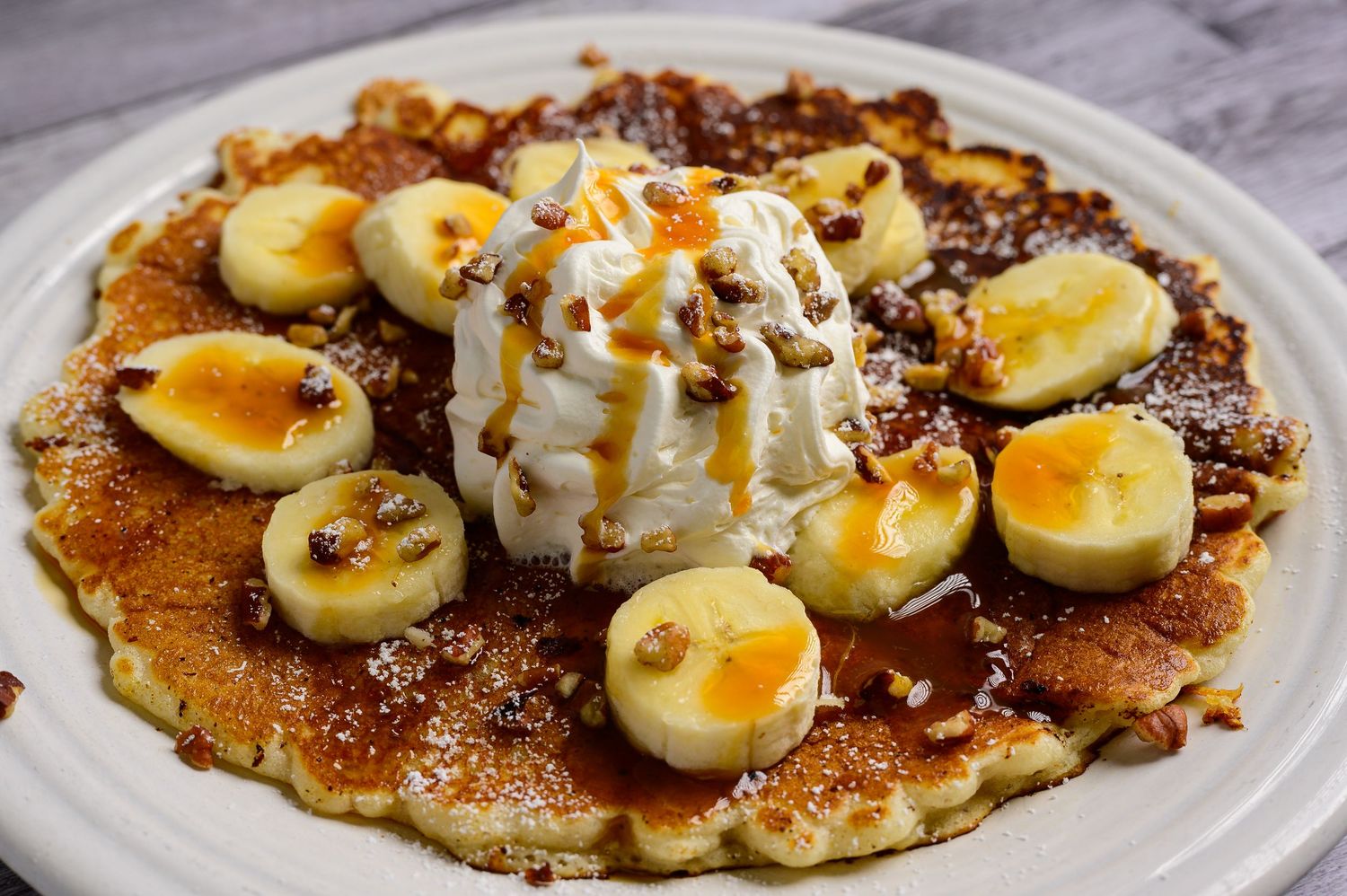 Banana foster pancake topped with whipped cream, caramel drizzle, banana slices, and chopped nuts on white plate.