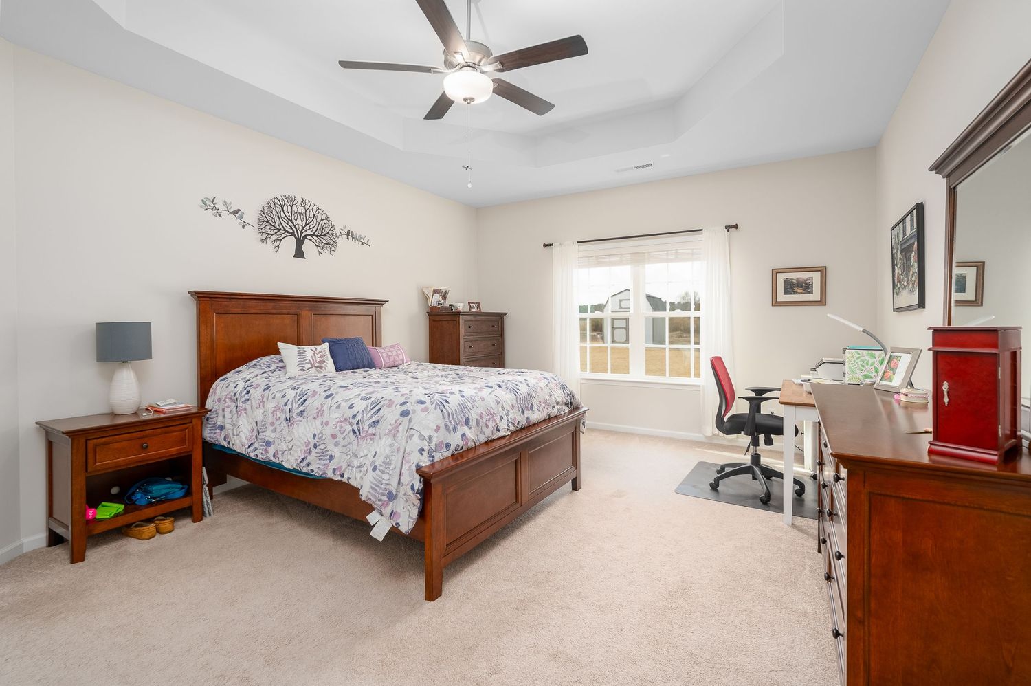 Spacious bedroom with tray ceiling, ceiling fan, wooden furniture, carpet, and a desk near the window.