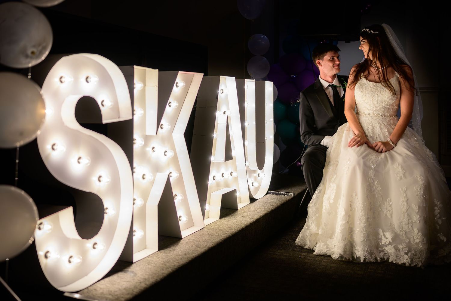 Newlyweds pose beside illuminated marquee letter sign SKAU at elegant wedding reception.