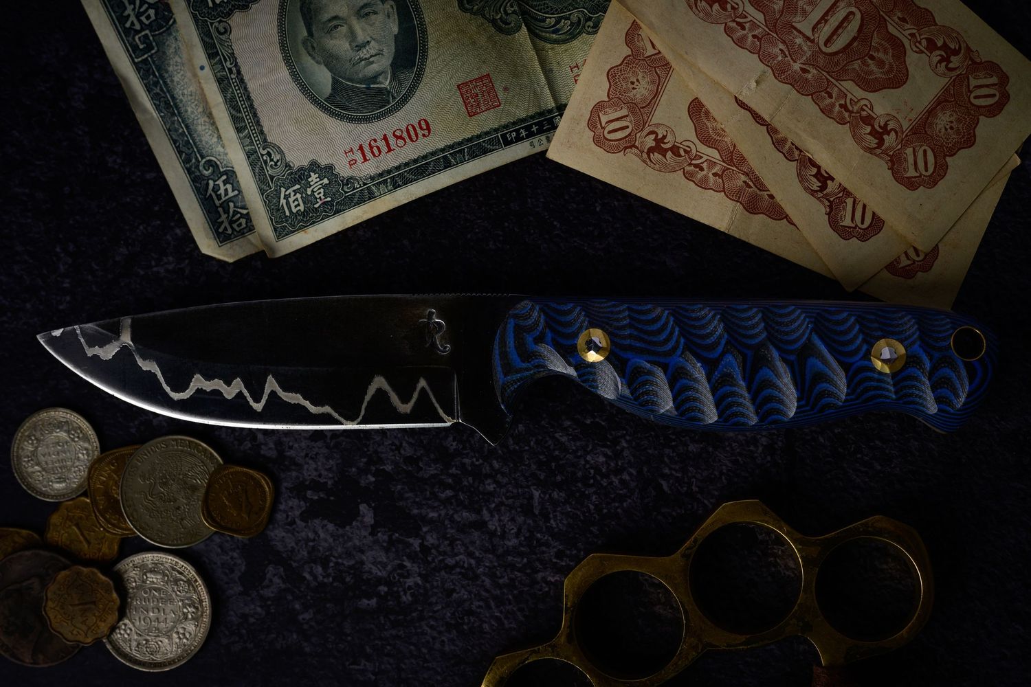 Handmade Damascus steel knife with blue handle beside vintage banknotes, coins, and brass knuckles.