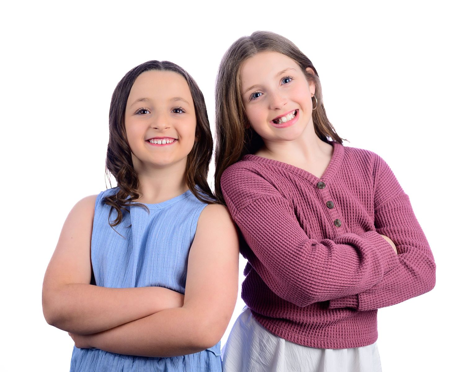 Two smiling young girls pose together, one in blue dress, one in pink sweater, arms crossed.